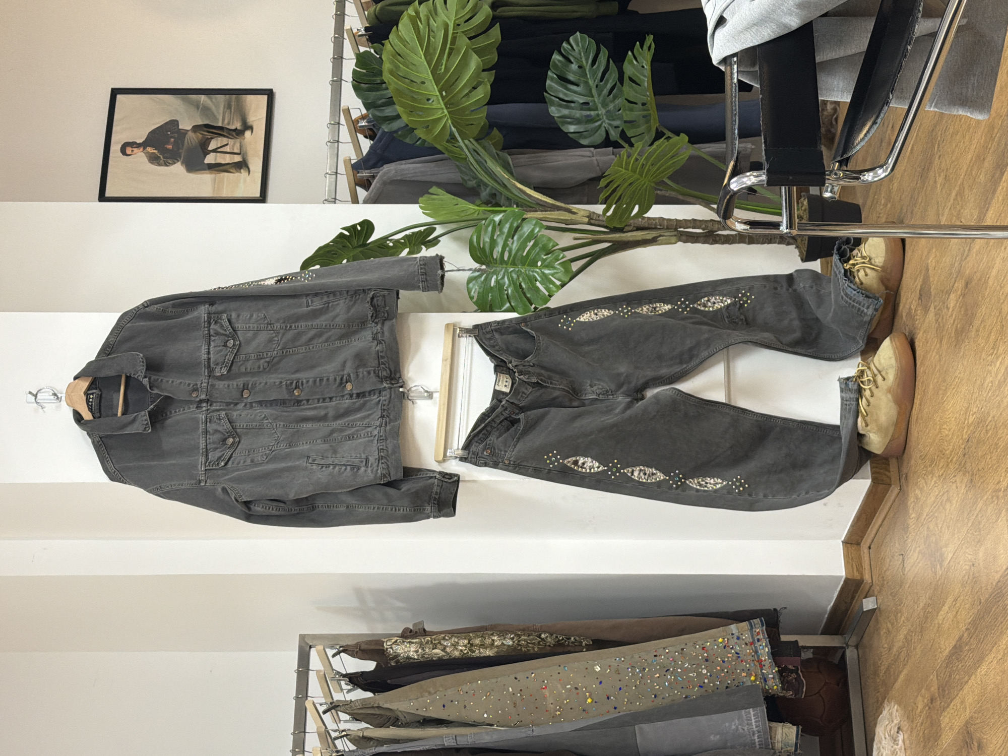The image depicts a fashion retail setting, likely a boutique in London, United Kingdom. A grey denim jacket and matching jeans with decorative embellishments are displayed on a white wall above a lower rack of clothing. A framed picture hangs above the ensemble. To the right, a faux Monstera plant and a partially visible black chair add to the scene. Two pairs of tan-colored shoes are positioned next to the jeans on the right. The environment suggests a curated, stylish retail space with a focus on unique or embellished apparel.