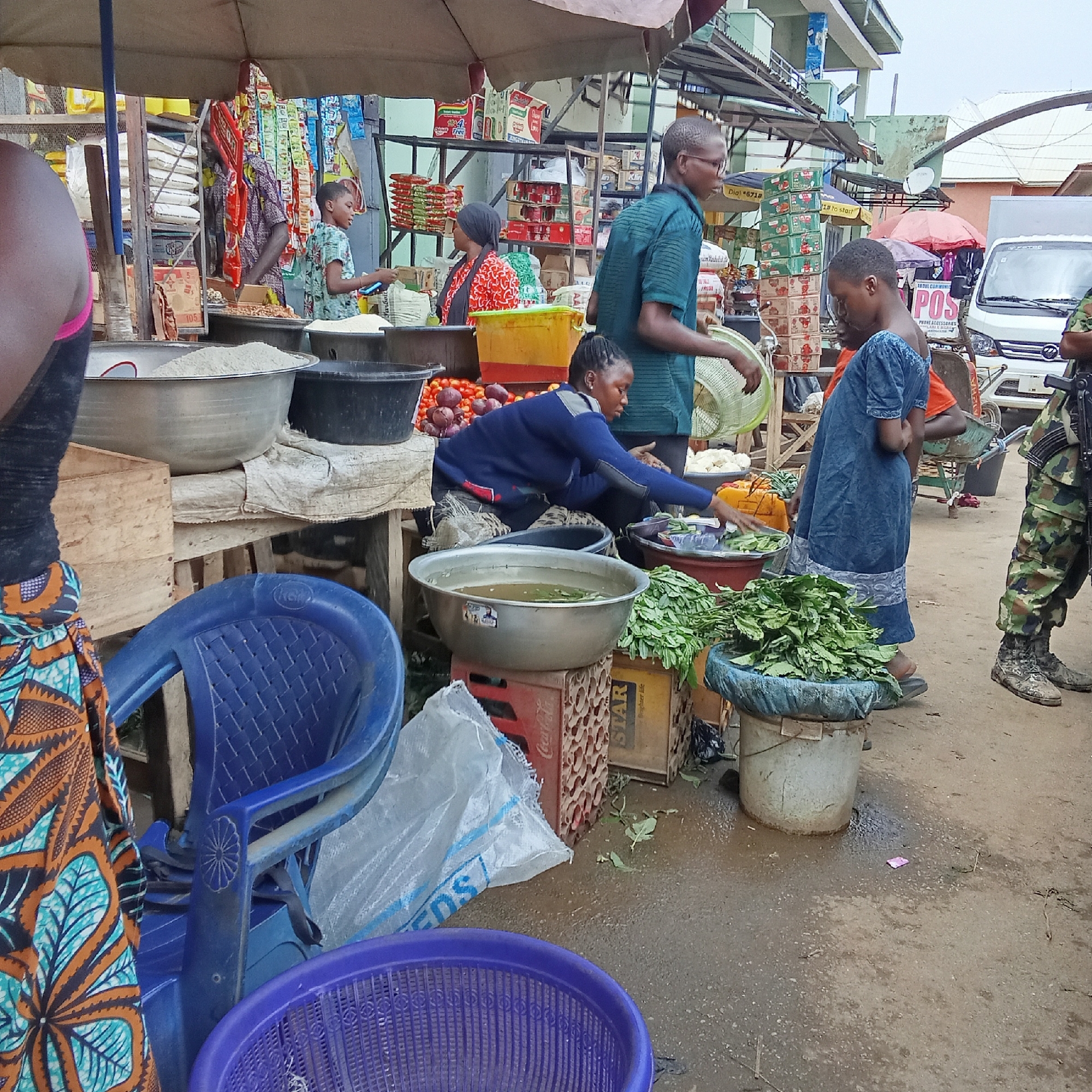 Here's a detailed description of the image:
**General Scene:** The image shows a bustling outdoor market scene, likely in Akwanga, Nigeria, given the location identified in the file name. The area seems to be a mix of stalls and vendors along a street.
**People:**
* Several individuals are present, including vendors and customers.
* One vendor is shown wearing a blue sweater and sorting through vegetables at her market stall.
* Customers are shown examining goods, including a person wearing a green shirt.
* A uniformed military or police officer with a rifle is on the periphery of the scene.
**Objects:**
* Market stalls are laden with various goods including rice, tomatoes, fresh leafy greens and vegetables.
* Produce is displayed in basins, buckets, and crates.
* Plastic chairs and baskets are scattered in the foreground.
* A van is parked in the background with "POS" signage.
**Actions and Interactions:**
* Vendors are actively serving customers and organizing their goods.
* Customers are browsing and potentially purchasing items.
**Environment:**
* The ground appears wet, suggesting it might have recently rained or been cleaned.
* The area is crowded, indicating a busy market day.
* The buildings are visible in the background.