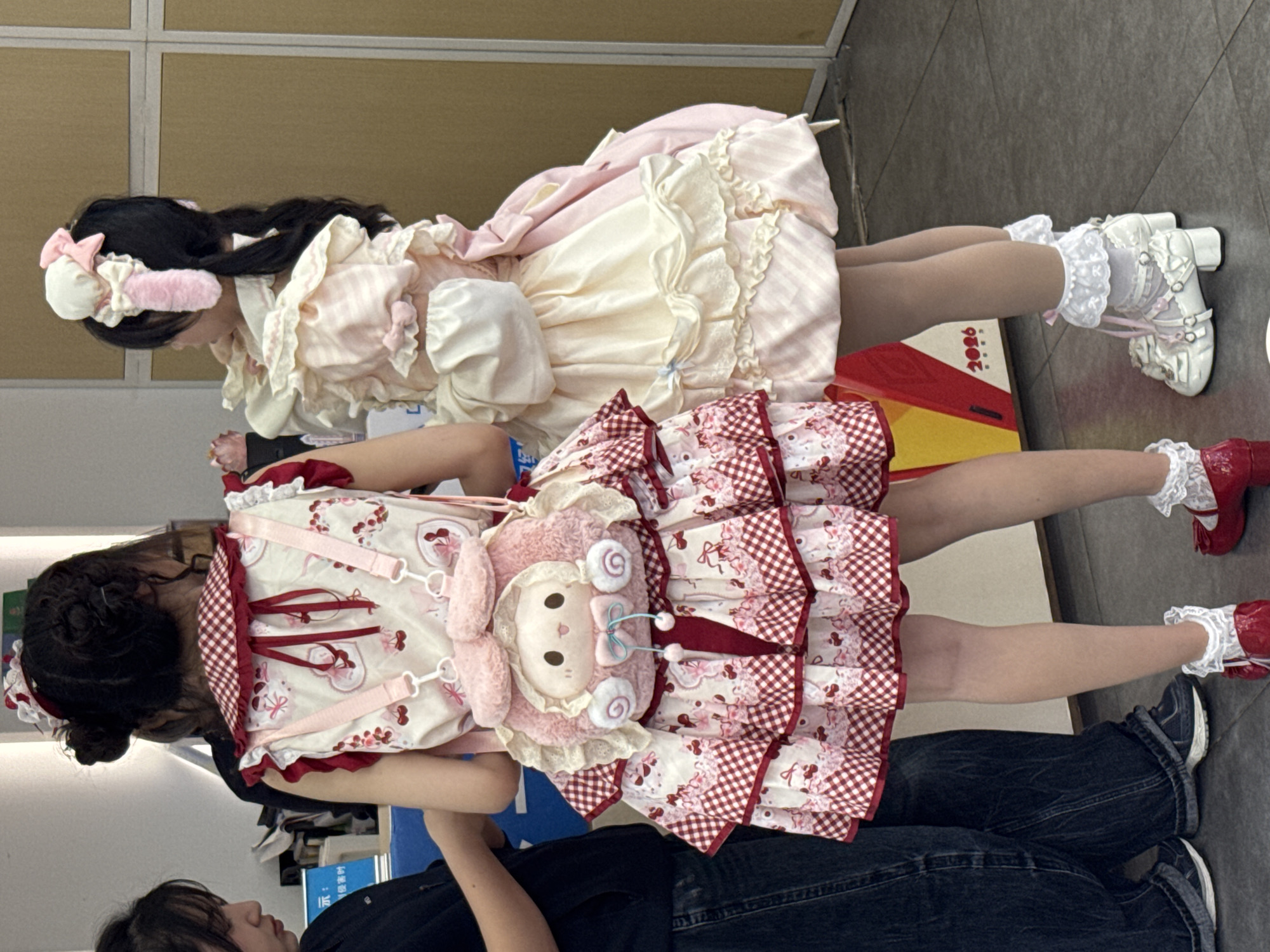 Two women are posing indoors, likely in Yuzhong District, China. The woman in the front is wearing a red and white lolita dress with a matching backpack in the shape of a lamb. The woman behind her is wearing a pink and cream-colored lolita dress with matching headwear. They both have frilly socks and lolita-style shoes. Another person is partially visible in the bottom of the frame. The background is bland.