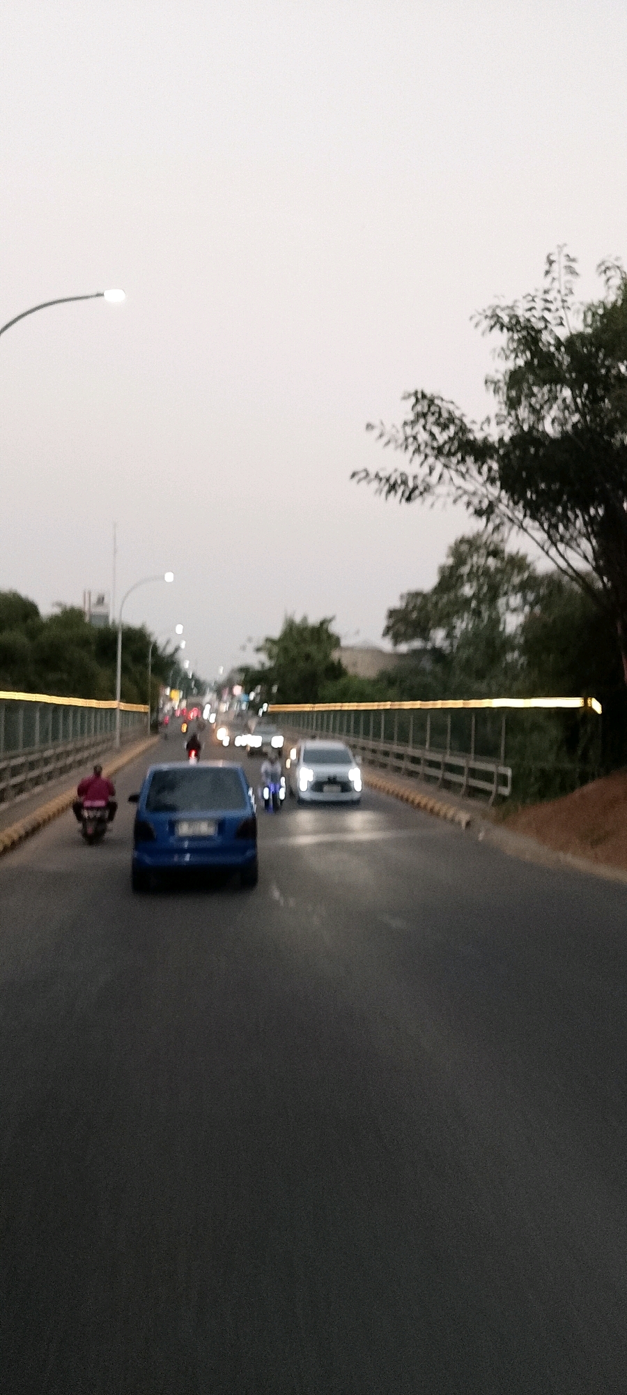 The image captures a scene on a road in Cileungsi, Indonesia, likely during dusk or a cloudy day given the low light conditions. The perspective is from a vehicle traveling on the road.
In the foreground, the asphalt road is visible. A blue hatchback car is the most prominent object, positioned in the left lane and moving away from the viewer. To the left of the blue car, a person on a motorcycle, wearing a pink shirt, is also traveling away from the viewer.
In the right lane, a white SUV is visible, also moving away. To the left of the white SUV and slightly ahead, another motorcycle with a rider, potentially with a glowing blue element on the wheels, is present.
The road appears to be elevated, possibly a bridge or overpass, as indicated by a railing with yellow accents running along both sides of the road. Trees and vegetation are visible on either side of the road, with more trees and foliage on the right.
Further down the road, the scene is blurred due to motion and distance, showing a line of streetlights and vehicle headlights, suggesting a moderately busy street. The sky is overcast, with a light gray hue. The overall impression is a typical urban or suburban street scene in Indonesia, characterized by common modes of transportation like cars and motorcycles.