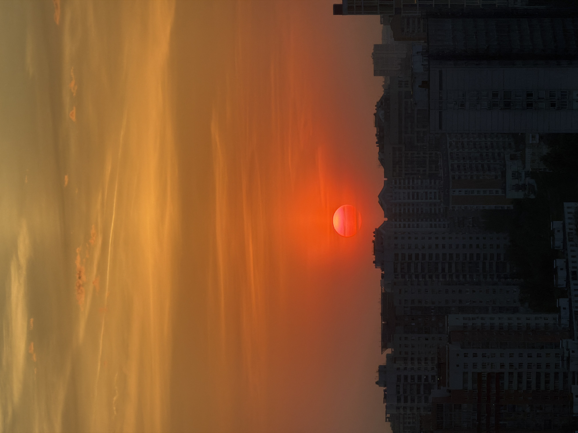 The image shows a vertical view of a cityscape during sunset or sunrise. On the right side, numerous high-rise buildings with multiple windows form a dense urban landscape, their appearance muted in tone. A bright red sun dominates the scene, positioned near the skyline. The sky, transitioning from orange near the sun to yellow further away, is partly obscured by thin, vertical clouds. The cityscape is purported to be in Khimki, Russia.