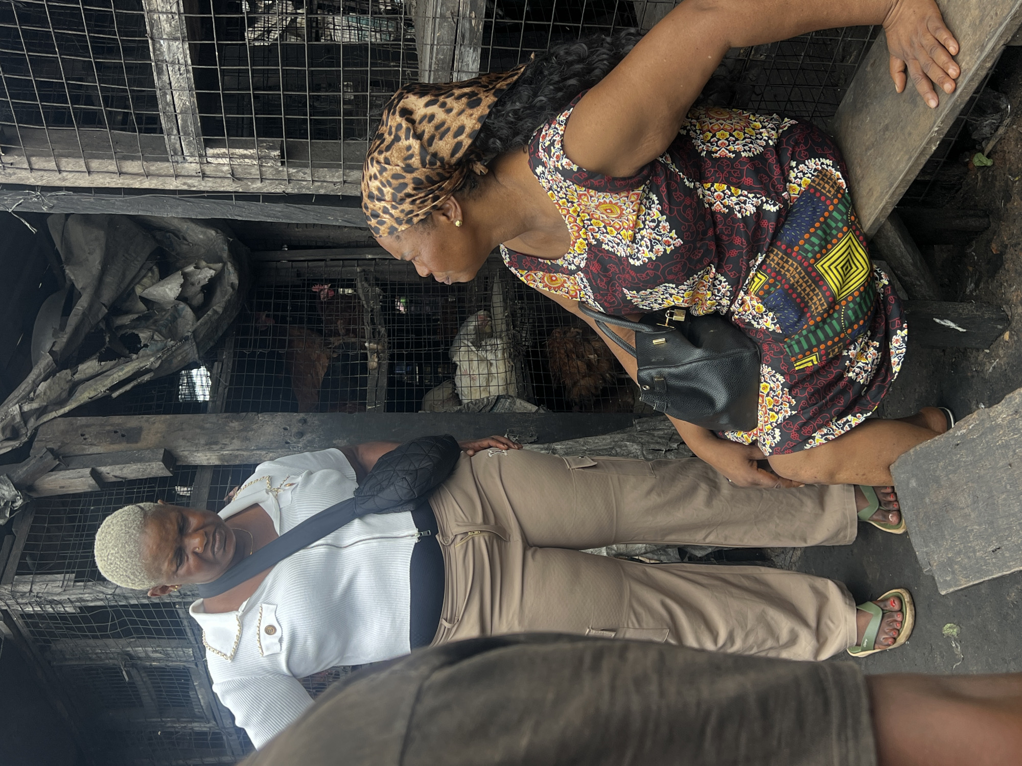 In Nkpor, Nigeria, two women are seen at what appears to be a market or animal enclosure. The woman in the foreground, with blonde, short-cropped hair, wears a white collared shirt and tan pants. She has a black cross-body bag slung over her shoulder. The second woman, positioned slightly behind and to the right, has her hair covered with a leopard-print scarf and wears a colorful, patterned dress. She also carries a black handbag.
Both women are interacting with the environment, which includes cages and what seem to be live chickens or other fowl within them. The scene suggests a commercial setting where live animals are sold. The lighting indicates it is daytime. The ground is concrete, and there are wooden structures and metal cages forming the stalls. The overall atmosphere is that of a busy market.