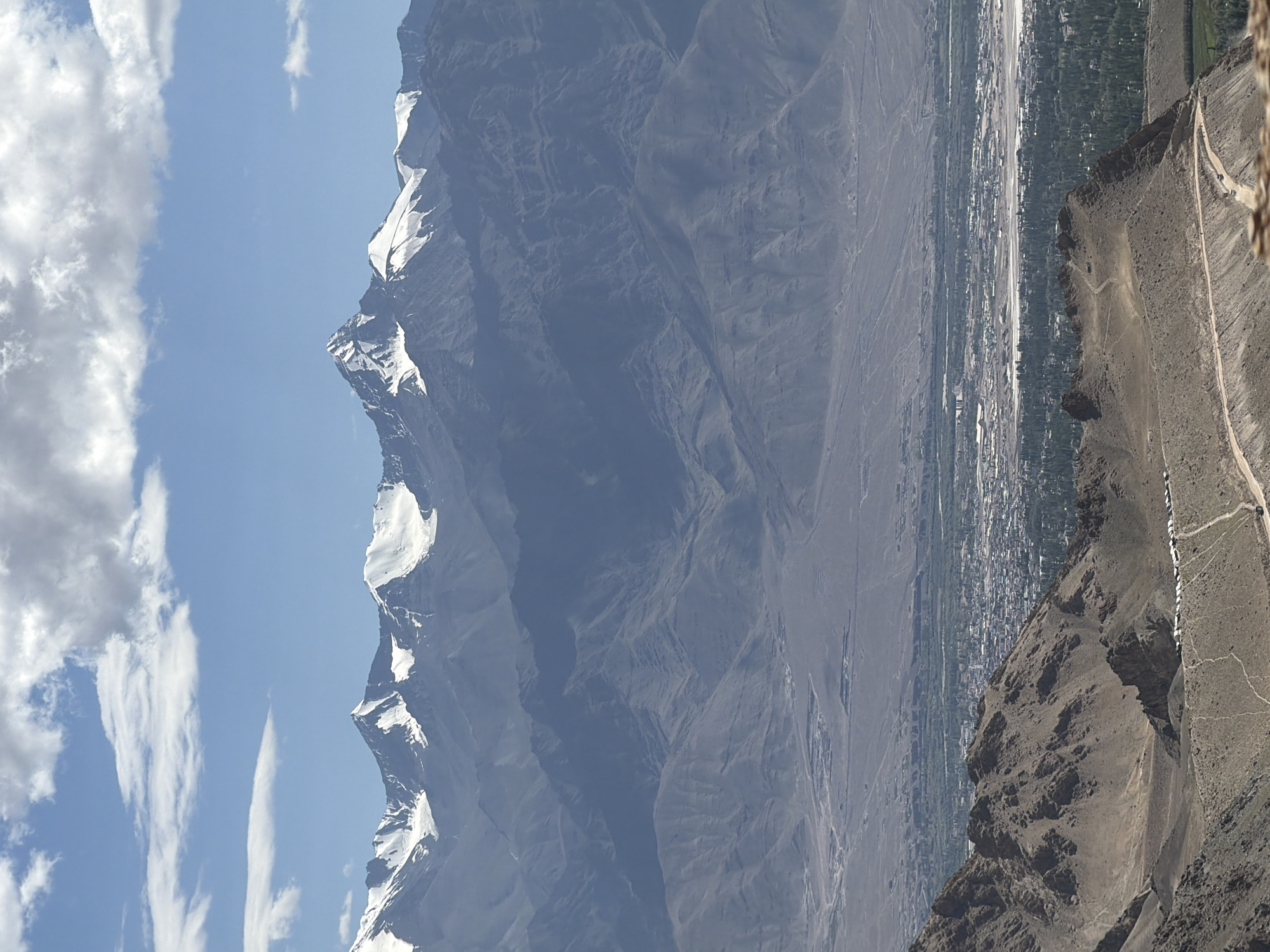 A wide-angle, high-angle shot captures a vast mountainous landscape under a partly cloudy blue sky. The foreground features rugged, rocky terrain with a dirt road winding through it, and a line of parked vehicles is visible along the road. In the midground, a city, identified as Leh, India, is spread out in a valley. The city appears dense with buildings and is surrounded by lush green vegetation. Further in the background, majestic snow-capped mountains dominate the horizon, their imposing peaks reaching towards the sky. The overall scene conveys a sense of natural grandeur and the presence of human civilization nestled within it.
