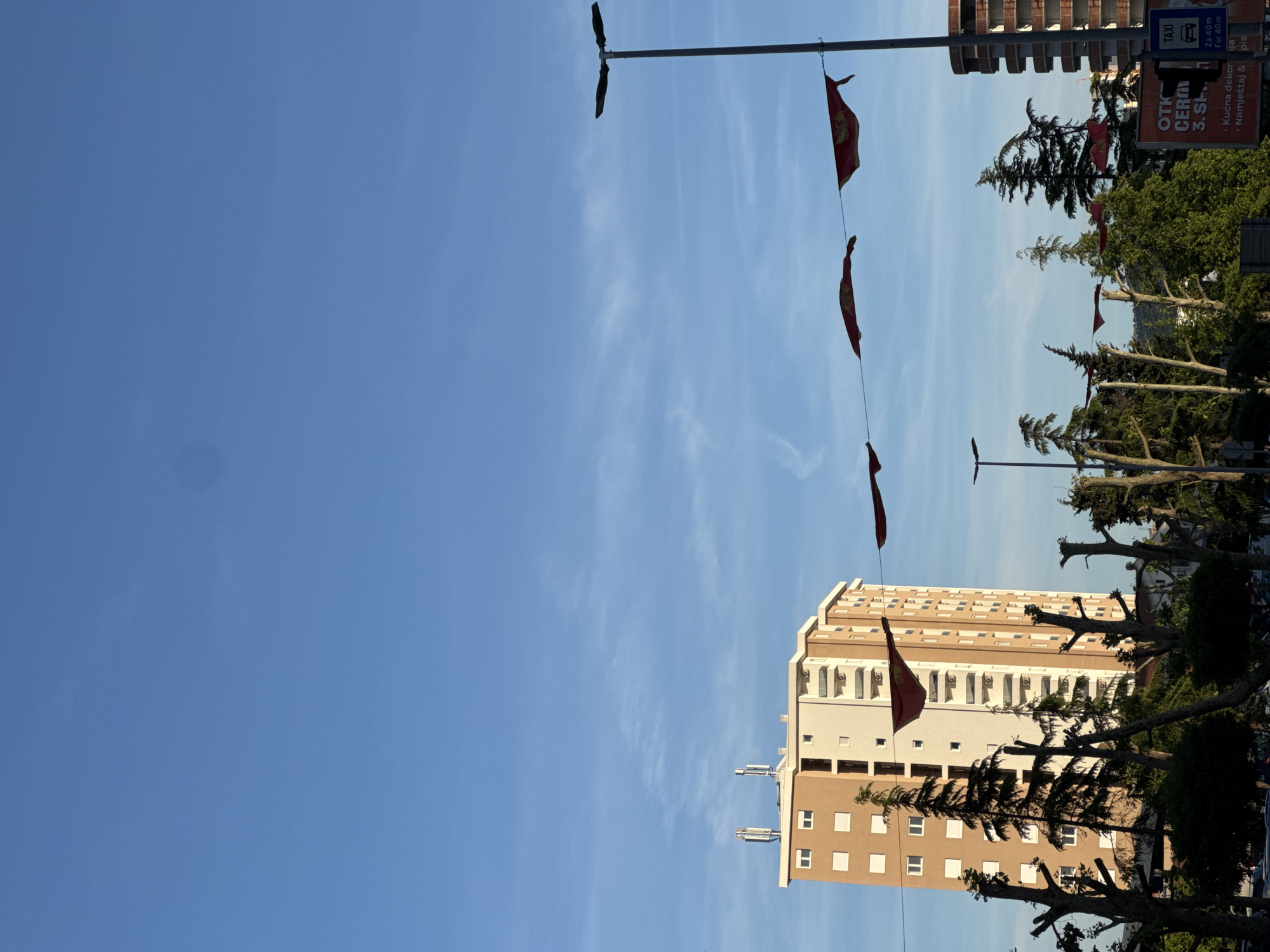 The image depicts a street scene in Bar, Montenegro, with a tall building, street lamps, and flags strung overhead. The sky is blue with wispy clouds. The building is tan and white with multiple windows and balconies. Trees with trimmed branches line the street. A sign on the right advertises a taxi service and a home decor store.