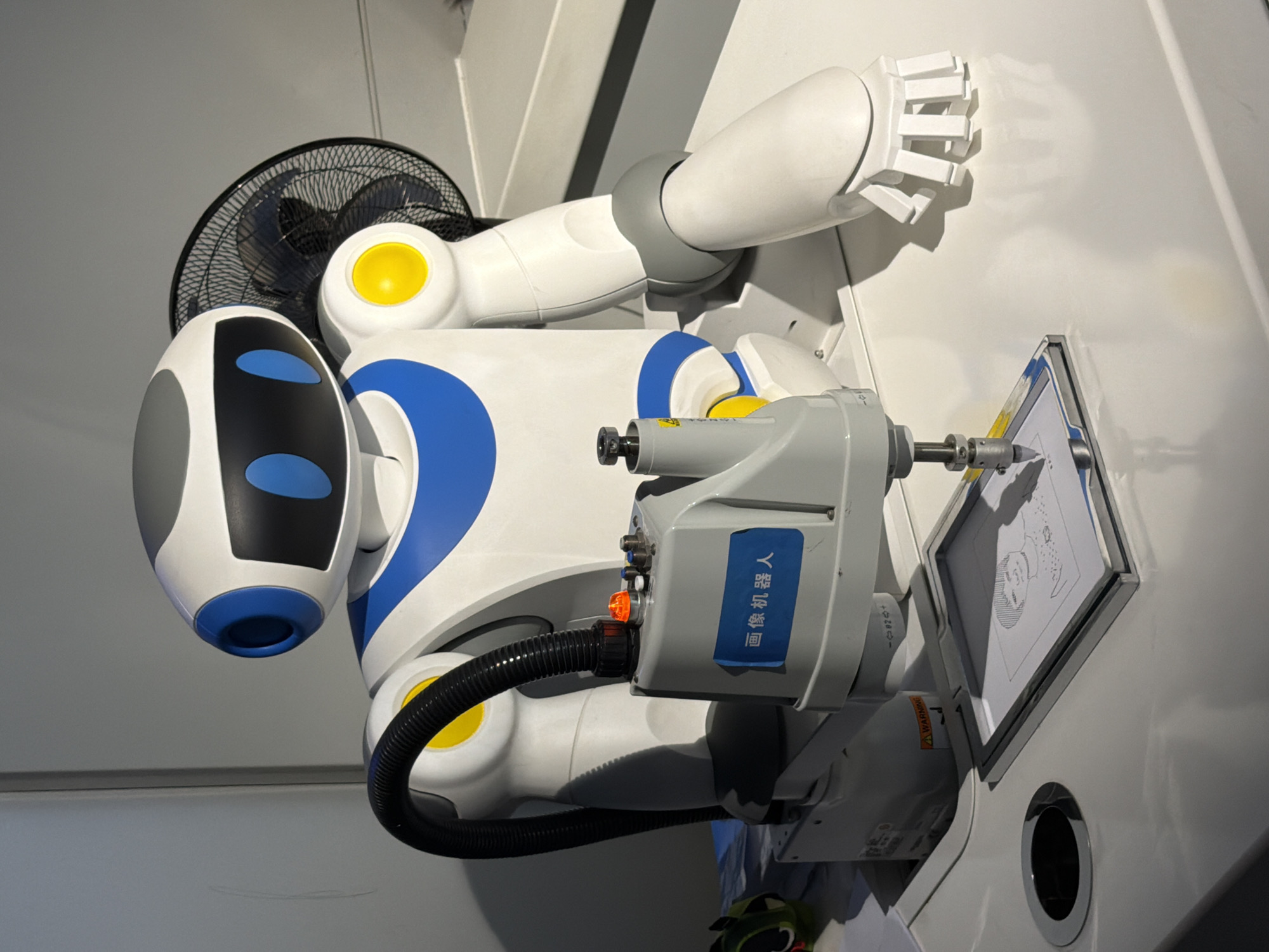 In a brightly lit indoor environment, a white and blue humanoid robot with a black head and blue circular eye accents is positioned on a white surface. A black corrugated tube connects from the robot's shoulder area to a gray mechanical arm, which holds a drawing tool over a framed piece of paper. The paper displays a partially drawn portrait. The robot has a yellow circular detail on its right shoulder. Behind the robot, a black oscillating fan is visible. The scene appears to be in a display or demonstration area. The location is described as 江北城街道, China.