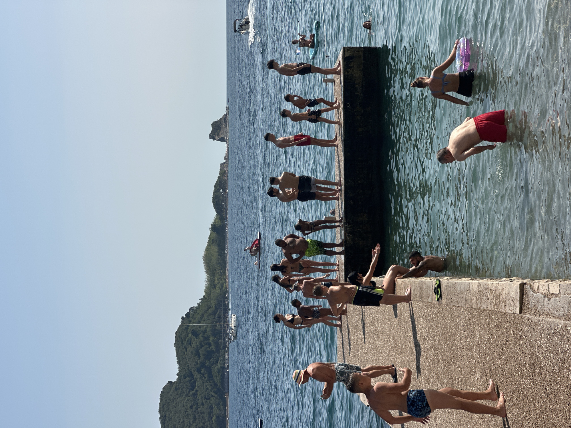 The image depicts a scene at a coastal location in Budva, Montenegro. A group of people are gathered on a concrete pier extending into the clear blue water. Many of the individuals are dressed in swimwear, suggesting a recreational activity like swimming or sunbathing. Several people are standing along the edge of the pier, appearing to be preparing to jump into the water, while others are already in the water, some with inflatable rings.
In the background, a boat is visible on the water, and a tree-covered hillside or island is present on the left side of the frame. The sun is shining brightly, casting shadows on the pier and illuminating the water, indicating it is likely daytime, possibly midday or afternoon. The atmosphere appears relaxed and summery. No text is visible in the image.