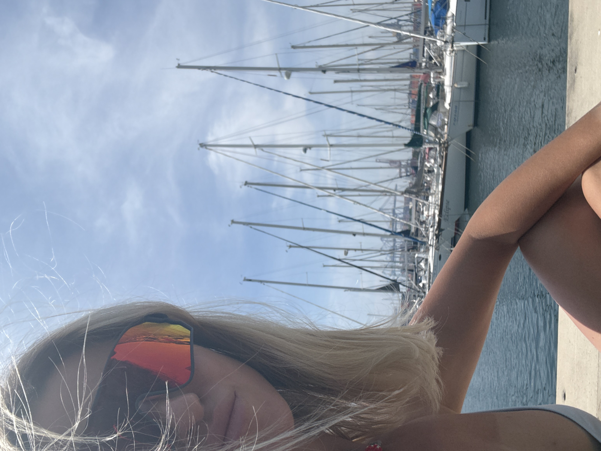 This image captures a close-up of a person relaxing by a marina in Valencia, Spain, on a bright, sunny day. The perspective is low, looking slightly upwards.
In the foreground, the lower portion of the frame is dominated by the upper body and head of a person with long, blonde hair. The individual is wearing large, sporty sunglasses with highly reflective orange and red lenses, which obscure their eyes and reflect the vibrant sky. Strands of blonde hair are scattered across the face and glasses, suggesting a gentle breeze. A bare shoulder and arm are visible, showing tanned skin, with the arms seemingly crossed or bent, resting on a concrete surface.
Behind the person, the scene opens up to a bustling marina filled with numerous sailboats. Their tall, white masts and intricate rigging create a dense, vertical pattern against the sky, extending into the distance. The boats are moored in calm, dark blue water, which gently reflects the light. A concrete dock or pier is visible at the edge of the water, where the person appears to be situated.
The sky above is a light blue with soft, wispy white clouds, indicating clear and pleasant weather. Bright sunlight illuminates the entire scene, creating strong highlights, particularly on the reflective sunglasses. On the hull of one of the sailboats, the name "Eclipse" is faintly discernible. The overall atmosphere conveys a sense of leisure and enjoyment in a beautiful maritime setting.