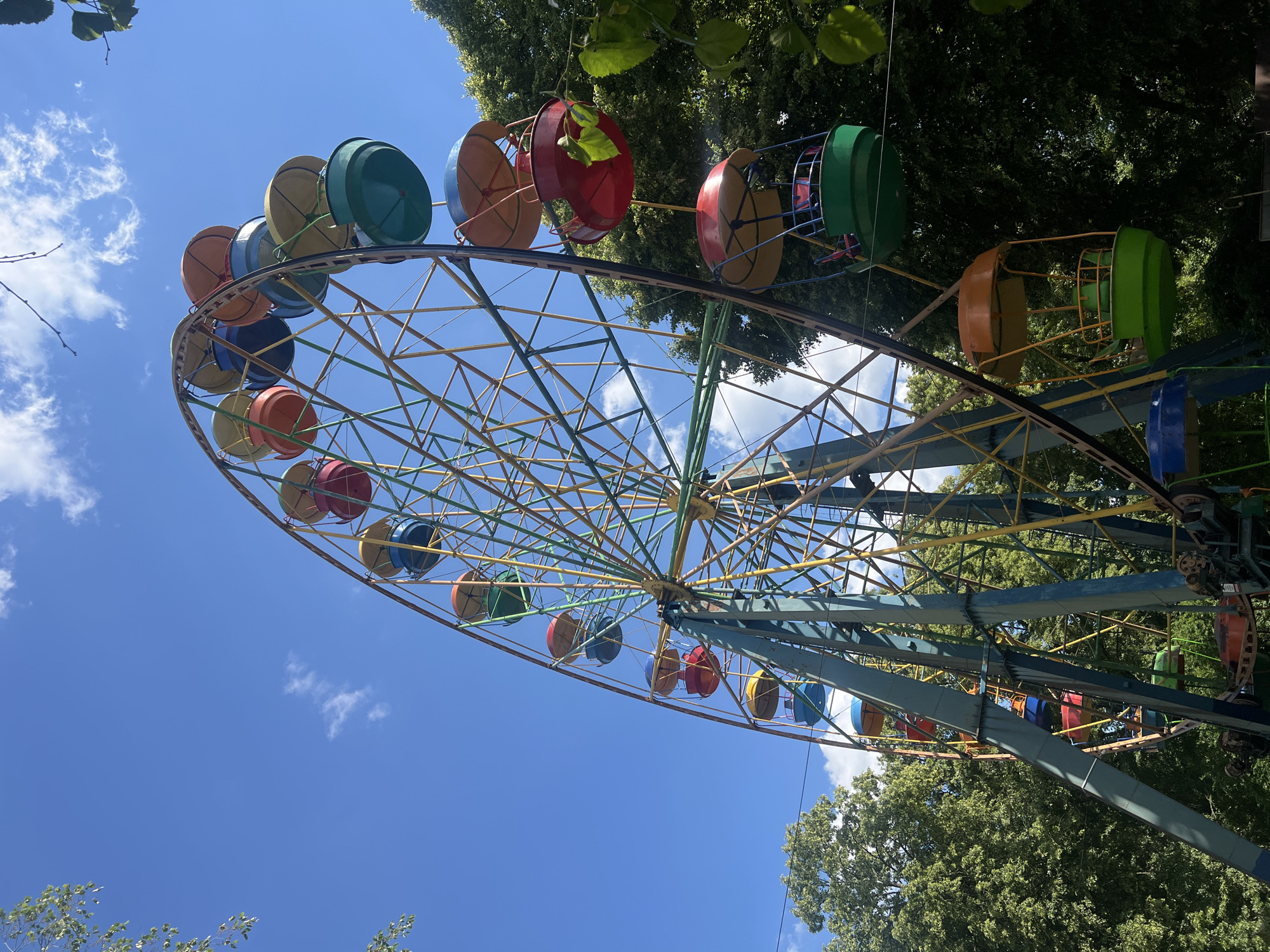 An outdoor, low-angle, full shot captures a vibrant, old-fashioned Ferris wheel against a bright blue sky with sparse clouds. The Ferris wheel is constructed of colorful metal beams in yellow, green, and blue, supporting a series of gondolas. These gondolas are also brightly colored, featuring red, blue, green, and yellow elements. The gondolas appear to be open-air, with simple seating inside. Dense green trees surround the base and rise behind the Ferris wheel, suggesting an park or amusement area. The overall impression is one of a sunny day at a classic amusement park, with a sense of nostalgia due to the slightly weathered appearance of the structure.