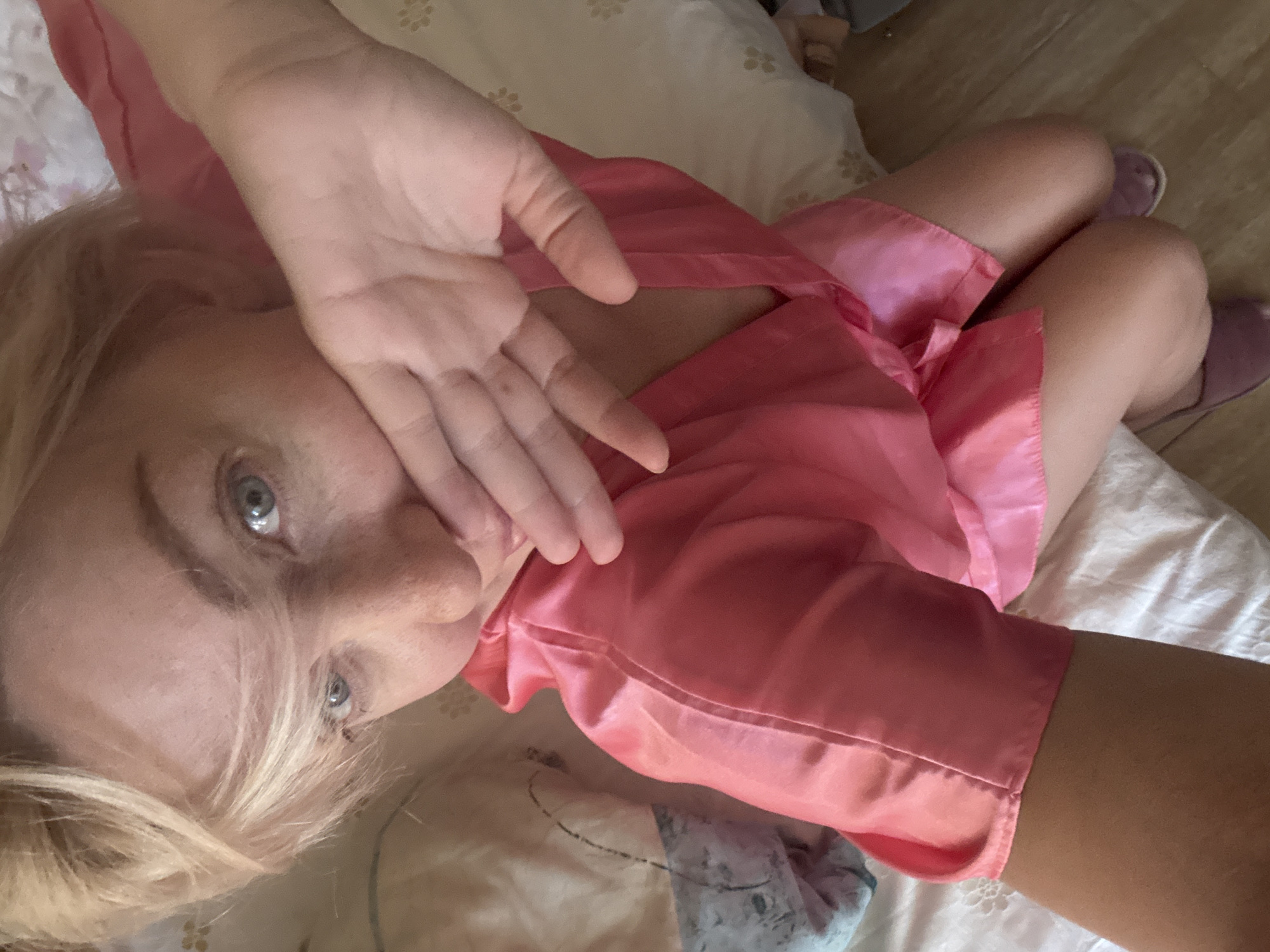 A blonde woman in a pink satin robe is lying on a bed. Her hand is covering her mouth. The room is dimly lit, with pillows and blankets visible. The floor is made of wood. There is no indication of the city of Sochi, Russia in this image.