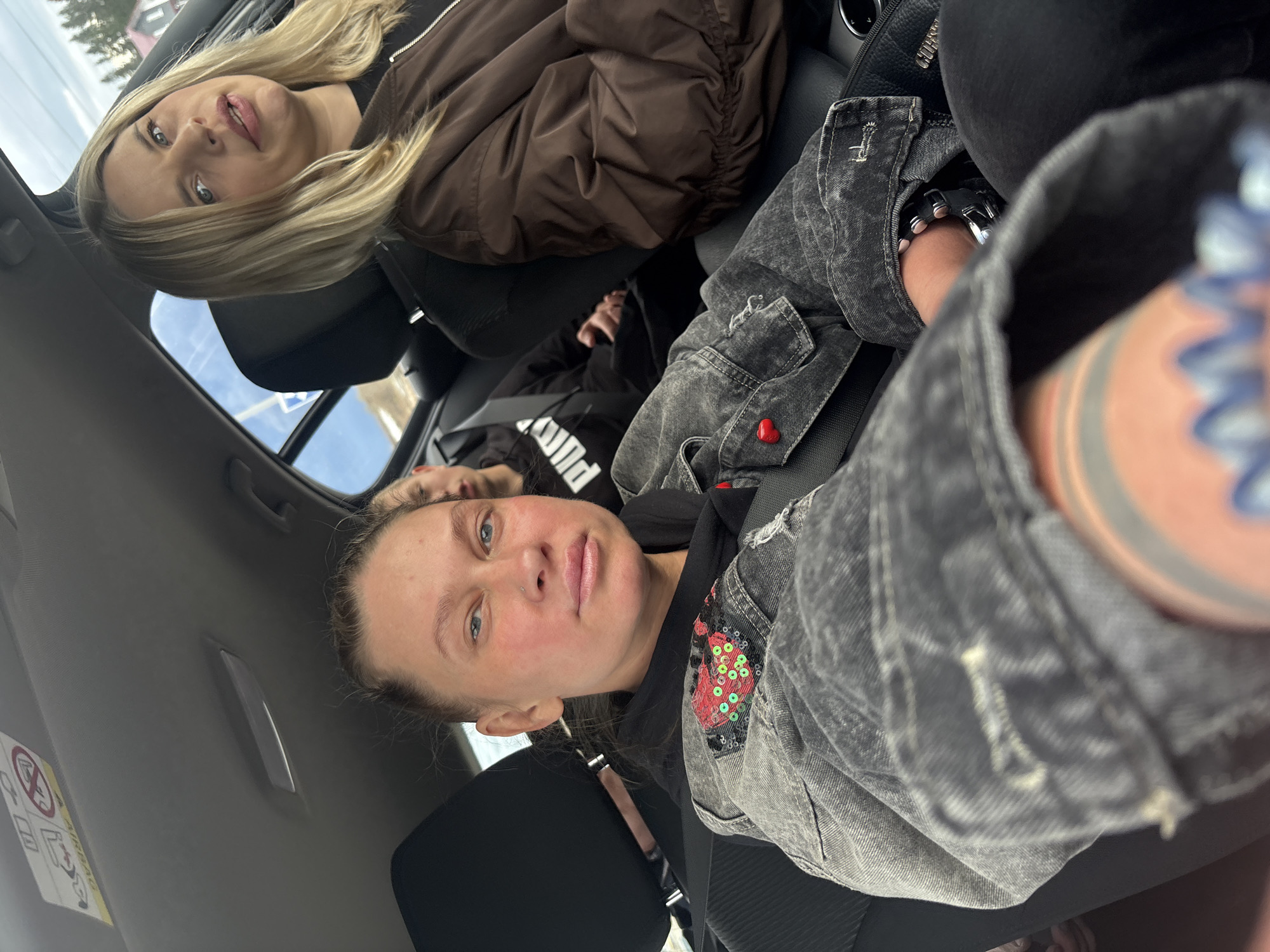 This image captures two young women inside a car, likely parked or in transit, with a third person partially visible between them. The photo is taken from a low angle, giving a dynamic perspective of the car's interior and its occupants.
In the foreground, a woman with dark hair, pulled back, looks directly at the camera. She has light blue eyes and a subtle nose piercing. She is wearing a black hoodie layered under a distressed grey denim jacket, which features a vibrant red and green sequined patch on the sleeve. A tattoo is visible on her right forearm/wrist, along with a metal watch or bracelet. Her expression is calm and neutral.
Behind her, in the middle passenger seat, another woman is partially visible, wearing a black top with white lettering, possibly reading "PUMA".
In the background, a blonde woman with long hair and light blue eyes also looks towards the camera, offering a faint smile. She is dressed in a brown puffer jacket over a black top. A black bag with a zipper is visible next to her.
The interior of the car is visible, including the seats, headrests, and part of the ceiling, which features an "AIRBAG" warning sticker. Through the car windows, an overcast sky is discernible, suggesting daytime, along with vague hints of buildings or trees outside. The overall mood is relaxed, with the subjects appearing comfortable within the vehicle. The scene is set in Kosoturikha, Russia.