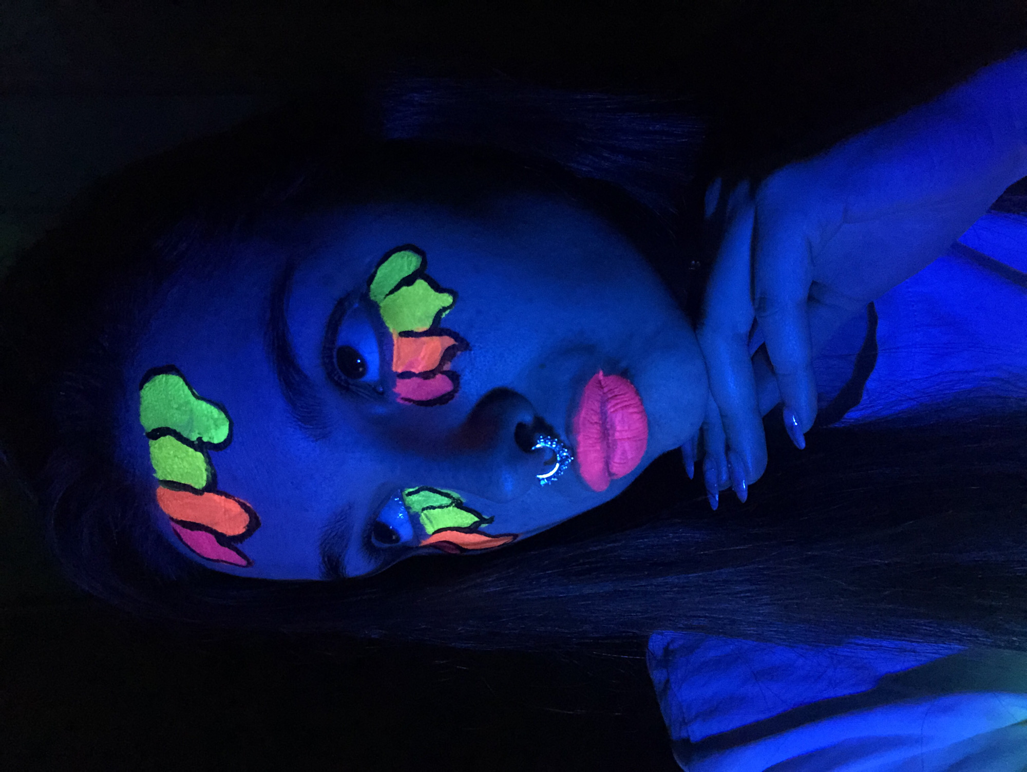 A person with dark hair and blue-lit skin is lying on their side. The person's face is adorned with bright, neon face paint. The paint is shaped like three stacked, rounded elements in green, orange, and pink. One stack is on the right side of the forehead and the other stack is under the right eye. The lips are painted pink. A silver-colored ring pierces the left nostril. The person's right hand is raised near their head, with long, manicured fingernails visible. The environment is dark, with the person illuminated by a blue light. The background is not visible. There is no evidence to suggest this image was taken in Noviy, Russia.