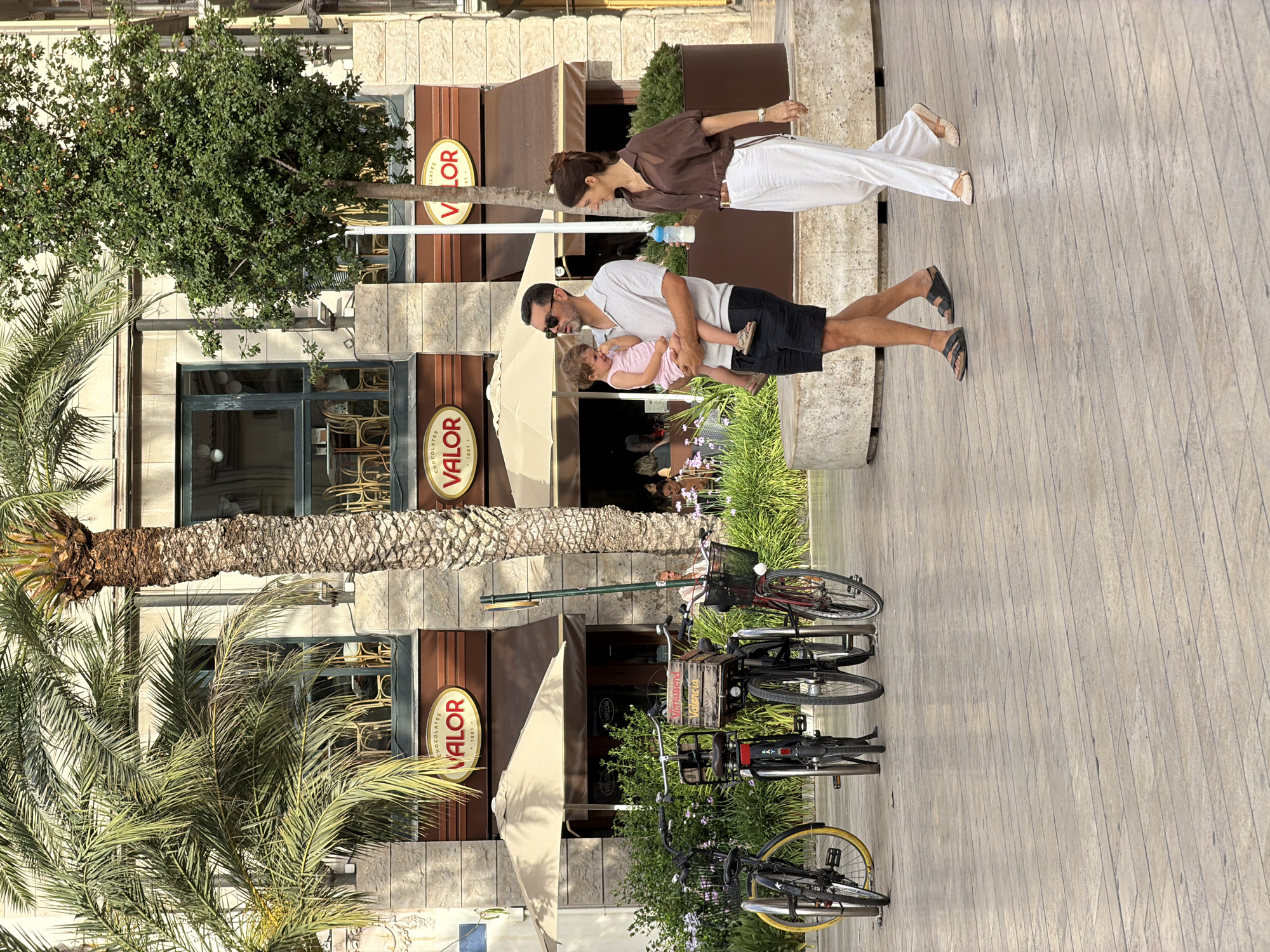 In an outdoor urban setting, a man carries a young child while walking alongside a woman. The man, wearing a grey polo shirt and black shorts, holds the crying child, who is dressed in a pink onesie. The woman, in a brown sheer blouse and white pants, holds a baby bottle. They are on a paved walkway with patterned wooden planks. Behind them, several bicycles are parked. In the background, a building with large windows and awnings features signage for "CHOCOLATES VALOR". Palm trees and a smaller leafy tree are visible, along with outdoor seating areas and umbrellas. The scene appears to be a sunny day.