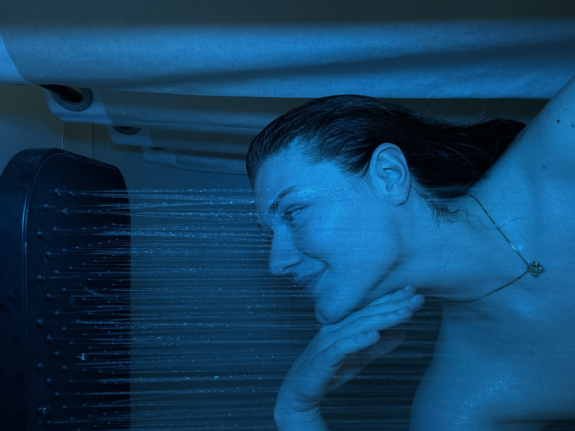 A person with wet dark hair, illuminated by blue light, is in a shower. The person's left hand is positioned near their face. A showerhead with multiple nozzles is emitting water streams. A necklace with a flower pendant is visible. The image appears to be taken indoors. No information suggests the location of Kazan, Russia, so this cannot be included.