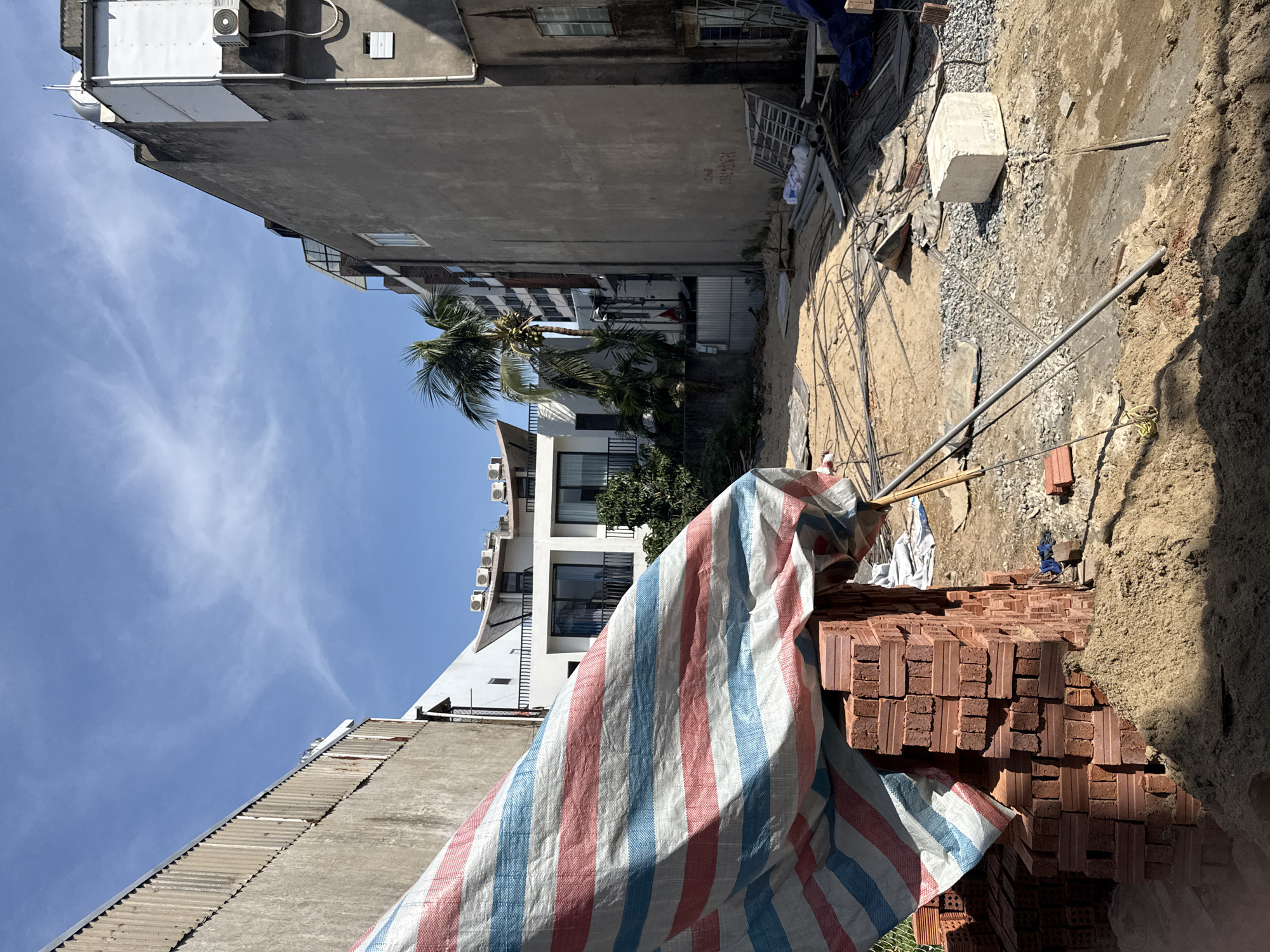 The image depicts an outdoor construction site or an undeveloped plot of land in an urban setting, likely in Da Nang, Vietnam, as indicated by the location context and visible text. The scene is bathed in bright daylight under a clear blue sky with scattered thin clouds, suggesting good weather, possibly midday or early afternoon.
In the foreground and mid-ground, substantial stacks of red bricks are prominent, partially covered by a protective tarp with distinct red, white, and blue stripes. The ground is uneven, consisting of loose sand, gravel, and packed earth, with various construction materials scattered across it, including rebar, metal pipes, and a large concrete block.
The plot is flanked by several buildings. On the left, a multi-story concrete building with a gray, unpainted facade and visible weathering stands tall. Air conditioning units are attached to its exterior. Faint black text, which appears to include "Đà Nẵng" and a phone number "0938426994", is visible on its wall. In the background, another multi-story white or light-colored building with multiple balconies and dark-framed windows adds to the urban backdrop. A tall palm tree, bearing coconuts, and other greenery are visible between these structures.
While no people are present, the collection of raw materials and the disturbed ground strongly imply ongoing construction or renovation activity. The overall impression is a bustling urban area undergoing development.