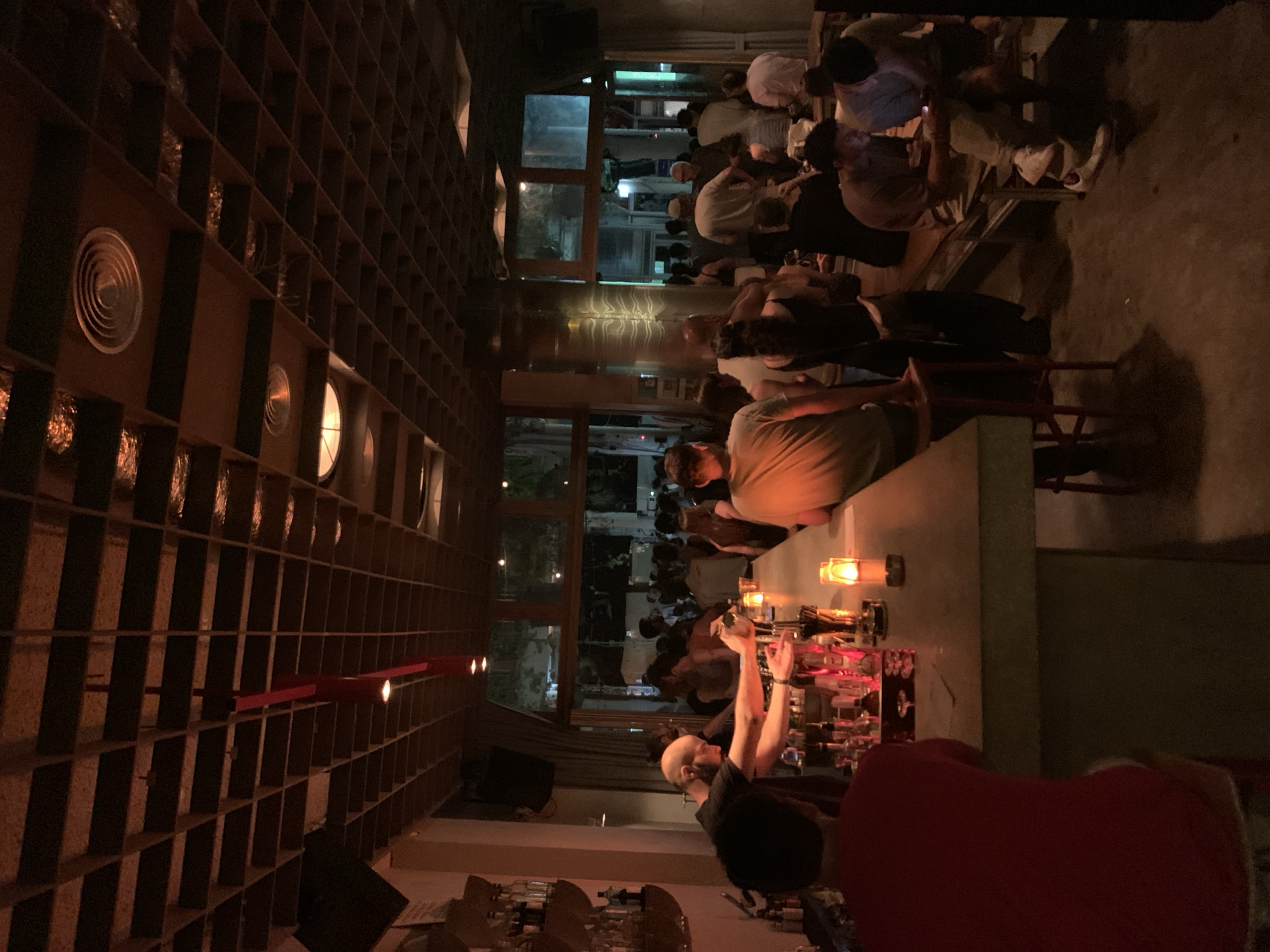 The image depicts the interior of a crowded bar or restaurant in Tel-Aviv, Israel, characterized by warm, ambient lighting. The ceiling features a distinctive geometric wooden grid structure with recessed lighting.
In the foreground, a person wearing a red shirt is seated at a concrete bar, facing away from the camera. A bartender, a bald man wearing a black t-shirt, is actively preparing drinks behind the bar, with bottles and bar tools visible. Several lit candles are placed on the bar counter.
The background shows a bustling scene with numerous patrons standing and mingling. Large windows and open doorways reveal an outdoor area, suggesting the establishment has an open-air component. The overall atmosphere appears lively and social. The architectural style and ambiance are consistent with trendy establishments found in cities like Tel-Aviv.