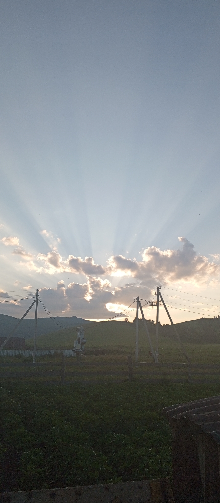 In the city of Khusainovo, Russia, a rural landscape is depicted at dusk. The sky is a pale blue, transitioning to a warmer hue near the horizon, where the sun is setting behind rolling hills. Beams of sunlight radiate upwards from behind scattered, fluffy clouds, creating a dramatic crepuscular effect. In the foreground, a field of dark green vegetation stretches across the lower portion of the frame. A wooden fence runs along the middle ground, separating the field from what appears to be a power substation with utility poles and wires. In the far left, a portion of a dark-roofed building is visible. The overall scene conveys a sense of peaceful, rural twilight.