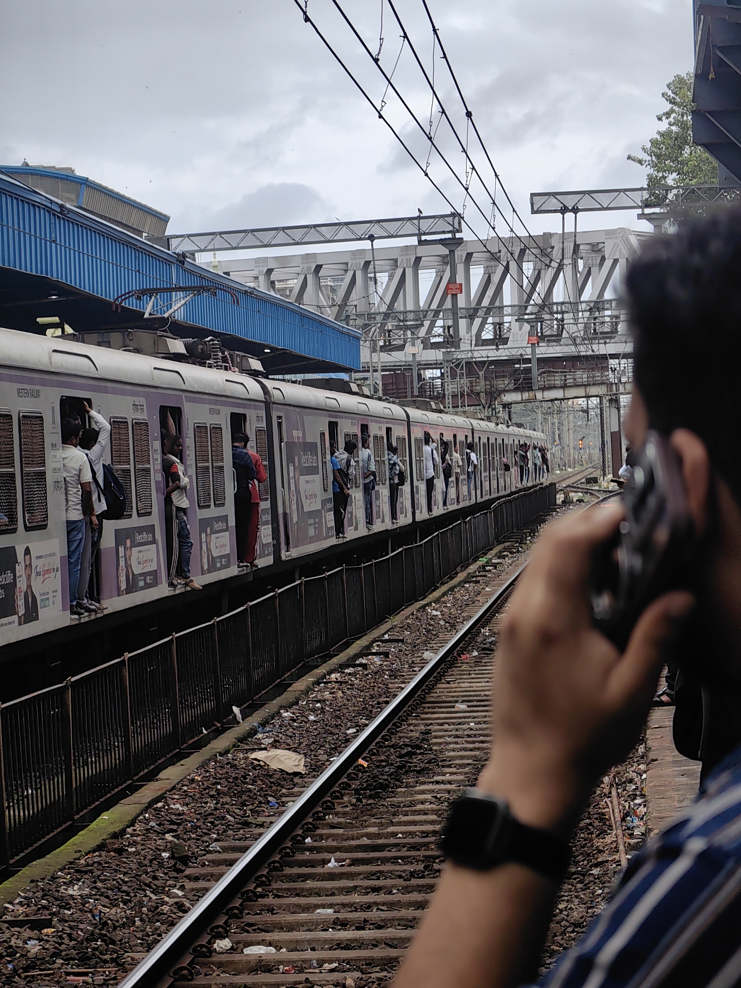 The image shows a commuter train stopped at a Mumbai suburban railway station, likely on the Western Railway line, as indicated by the signage on the train. The train is packed with passengers, many of whom are leaning out of the windows or standing on the exterior, a common sight during peak hours in Mumbai. The station platform has a blue elevated structure overhead, and in the background, a complex railway bridge or overhead structure is visible. The train tracks are lined with ballast, and some litter is present on the ground. In the foreground, a person is partially visible, holding a mobile phone to their ear, with their arm and watch also in frame. The sky is overcast with clouds.