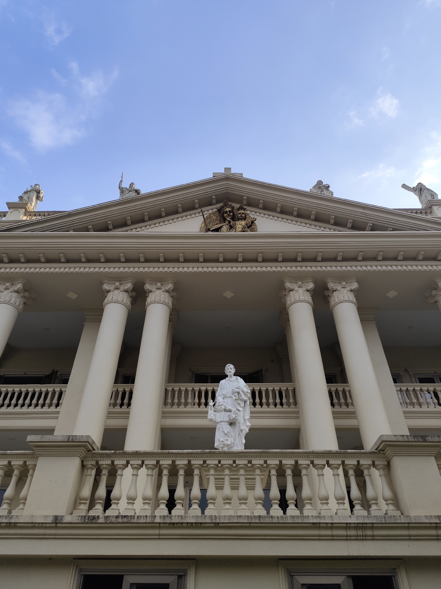 The image shows a low-angle view of a multi-story building with classical architectural elements. The building is light beige in color, featuring white columns with ornate capitals supporting the roof. A statue of a man and child stands on the balcony of the first floor. The building's roof is decorated with statues, including one in the center depicting two figures. Above the building, the sky is visible with scattered clouds. The building is located in Da Lat, Vietnam.