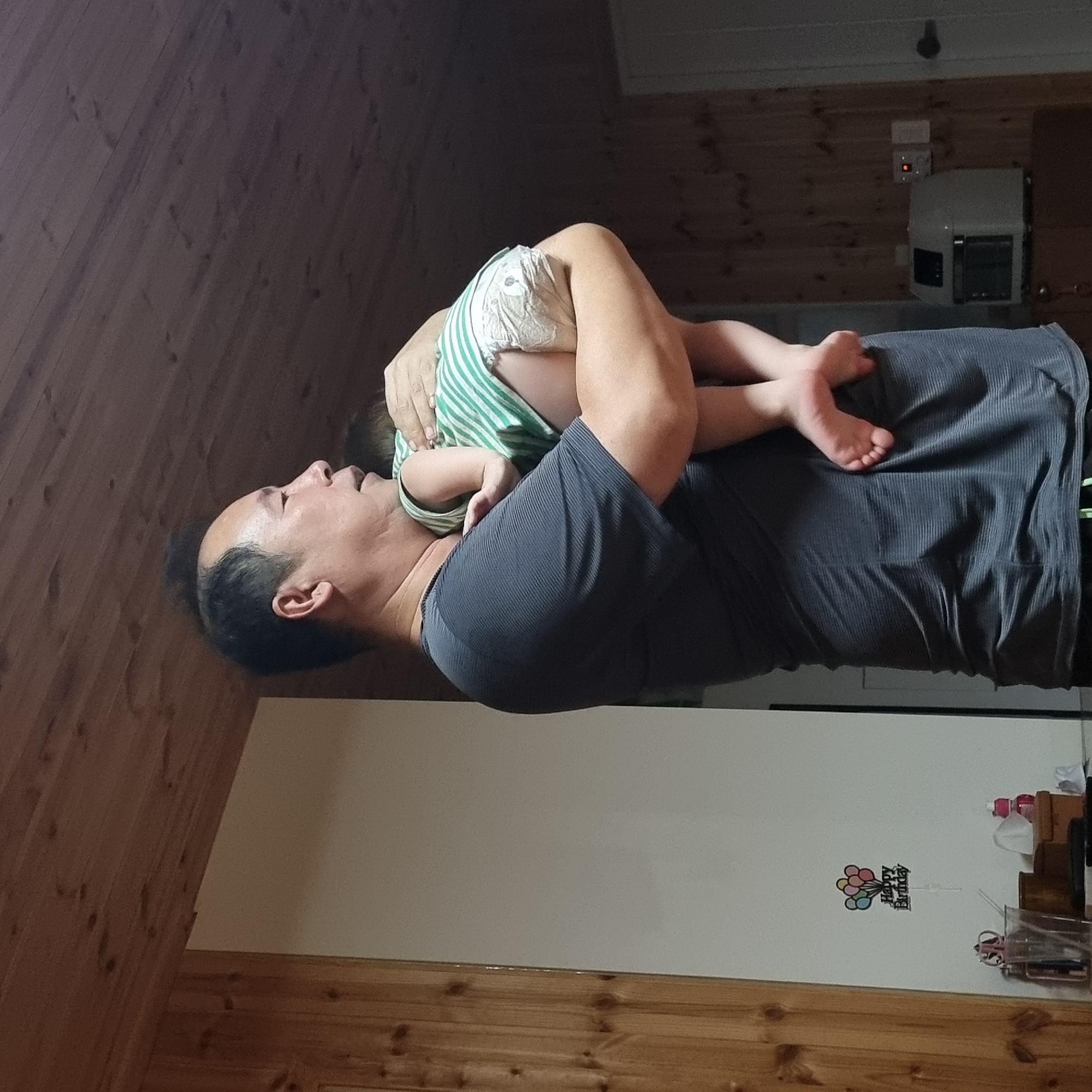 A man holds a baby wrapped in a green and white striped outfit and a diaper. The man is wearing a dark grey, striped t-shirt. The baby's feet are visible. A "Happy Birthday" decoration with balloons is on a white wall in the background. The room has wooden walls and a wood-paneled ceiling. There is a white appliance and a door visible in the background. The scene appears to be indoors.