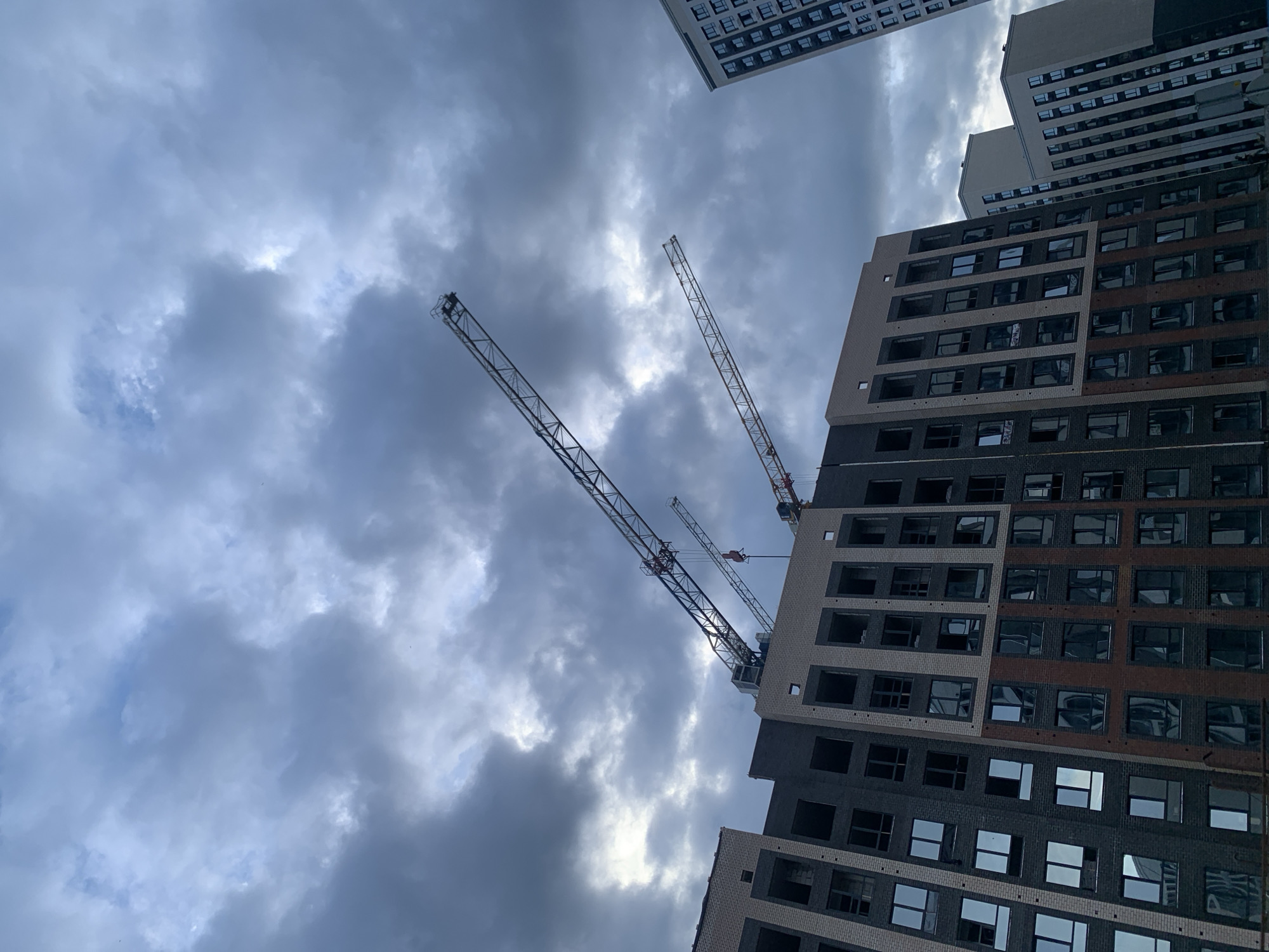 This is a photograph taken from below, looking up at a cloudy, overcast sky over a construction site in Stavropol, Russia. In the foreground, an unfinished apartment building is visible, featuring a combination of dark gray and light beige brickwork, with many windows either empty or reflecting the sky. Two construction cranes are prominently positioned, one larger and one smaller, silhouetted against the cloudy sky. To the right, another taller, more finished apartment building is visible. The overall scene suggests active construction in an urban environment under a somber sky.
