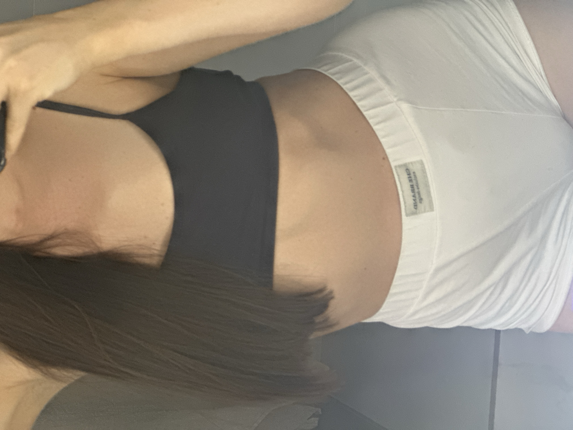 The image shows a close-up of a person's torso from the chest down to the upper thighs. The person is wearing a dark gray sports bra and white underwear. The underwear has a small tag with text on it. The person's skin is visible, and their hair is long and dark brown, falling over their shoulder. The background appears to be a plain wall, possibly in a bathroom, with some reflections suggesting a mirror. There is no visible indication of the city of Estachi, Russia, in the image.