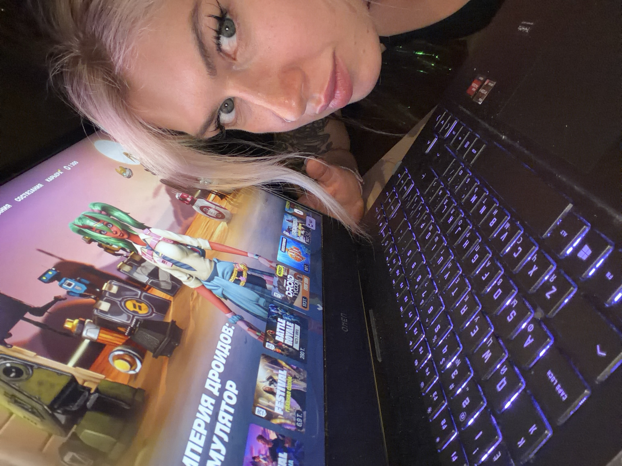 A woman with pale hair is looking at the camera while lying next to a laptop. The laptop screen displays the game "Fortnite" with the game selection menu visible. The keyboard of the laptop is lit up with a blue backlight. The laptop has an "OMEN" logo and also has AMD Radeon branding. The language displayed on the laptop screen is Russian.