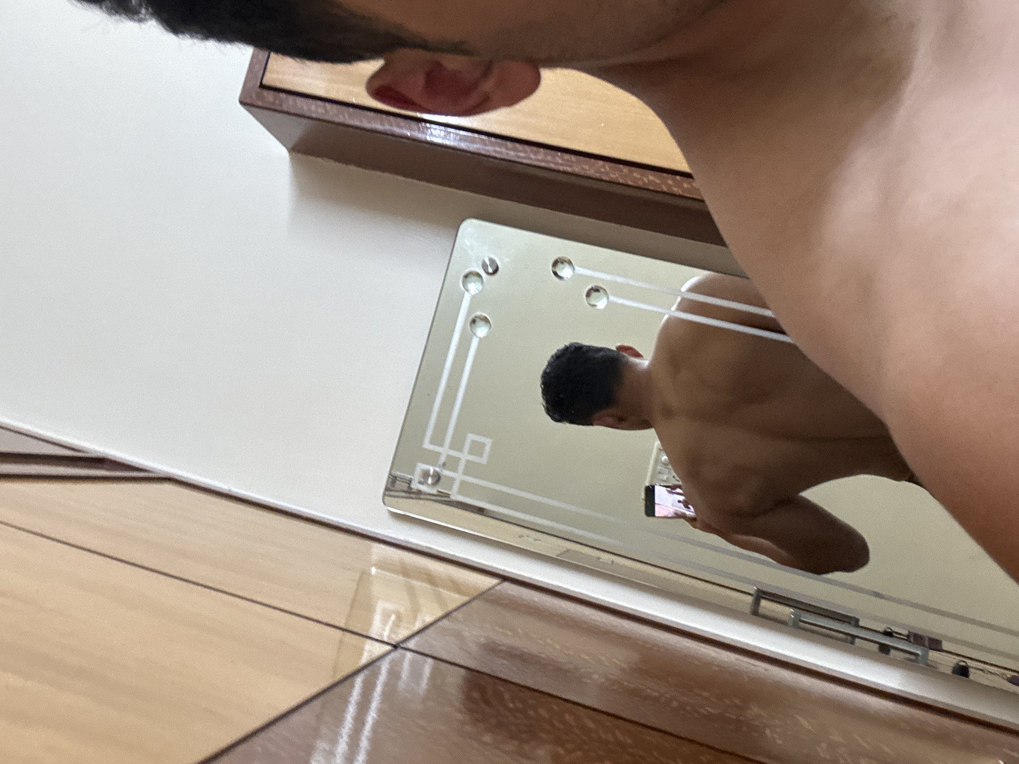 The image shows a person taking a shirtless selfie in a mirror. The person's face is partially visible in the upper left corner. The mirror is square-shaped with decorative elements and is mounted on a wall. Below the mirror is a wooden floor. The reflection in the mirror reveals the person holding a phone and taking the photo. The location is possibly Kolkata, India, but that cannot be definitively confirmed from the visual information.