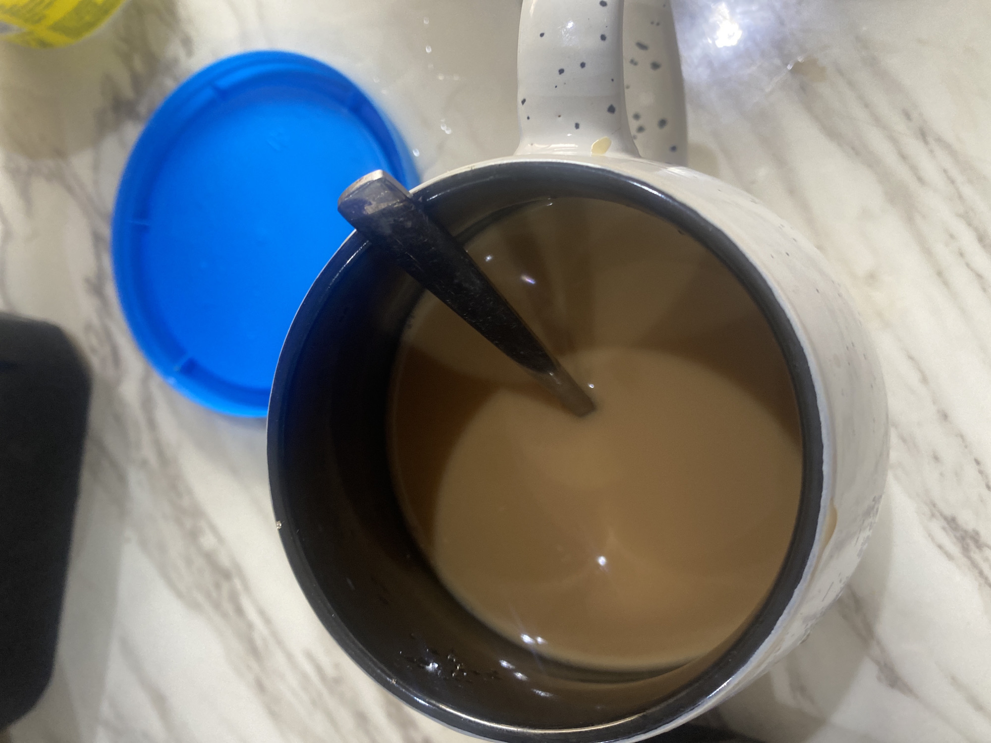 The image shows a top-down view of a coffee mug filled with coffee. A spoon is inside the mug, stirring the coffee. Beside the mug is a blue lid, presumably for a container that the coffee came from. The image is taken indoors and the background is a white countertop with a marbled pattern. There are no visible people. Given the lack of context within the image, it is impossible to determine a specific location in Lagos, Nigeria.