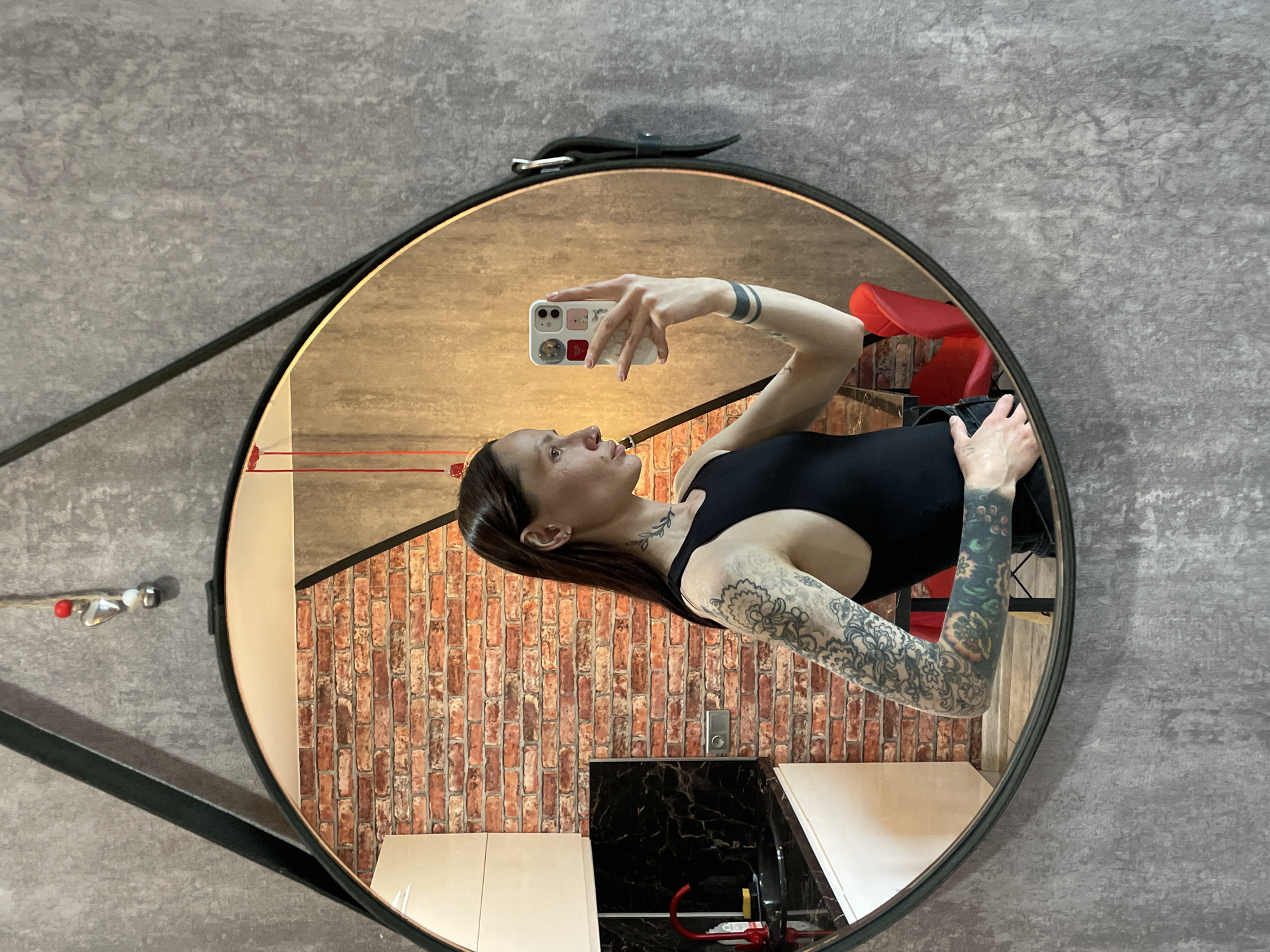 A reflection of a woman taking a selfie in a round mirror. The woman is wearing a black tank top and has tattoos on her arms and neck. The background of the reflection includes a brick wall, a black and white countertop, and a red chair. The mirror is hanging on a gray wall. The location may be Izhevsk, Russia, but this cannot be verified from the image alone.