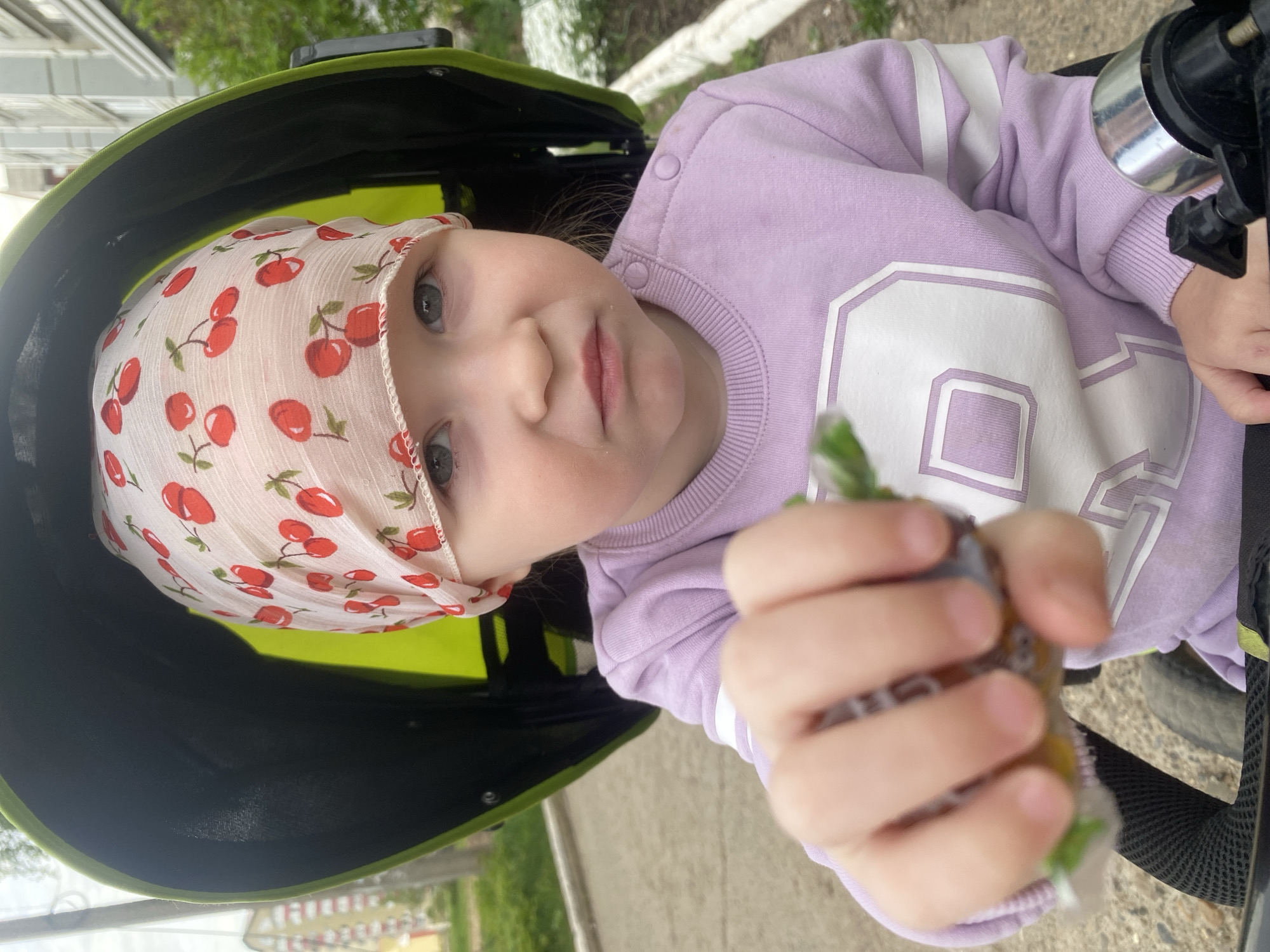 A young child with fair skin and gray eyes, wearing a light purple long-sleeved shirt with a white number "9" on the chest and a white headscarf with a red cherry pattern, is sitting in a green stroller. The child is holding a food pouch in their hand. The scene appears to be outdoors, potentially in the city of Chaikovsky, Russia, based on the context. A paved surface is visible near the stroller.