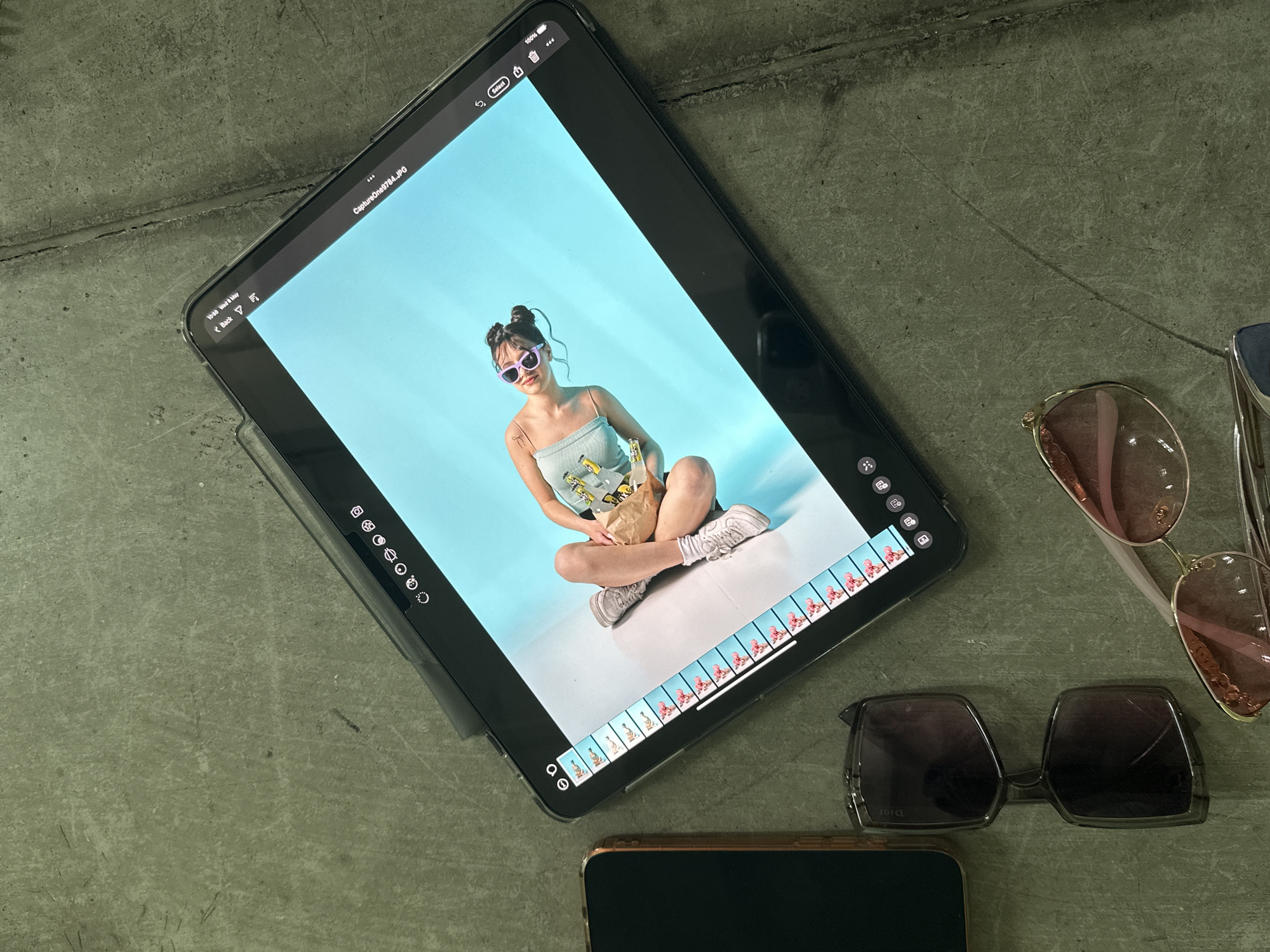 The image depicts an overhead view of a scene set against a dark gray surface. A black iPad is centrally positioned displaying an image of a woman with her hair styled in buns, wearing purple sunglasses and a light blue top, holding what appears to be a bag of chips or similar item. The woman is sitting in a cross-legged position against a light blue background.
Several pairs of sunglasses, including one with dark lenses bearing the inscription "Dior", are situated to the right of the iPad. A smartphone with a black screen is placed near the bottom edge of the frame.
The exact location in Yerevan, Armenia cannot be determined from this image.