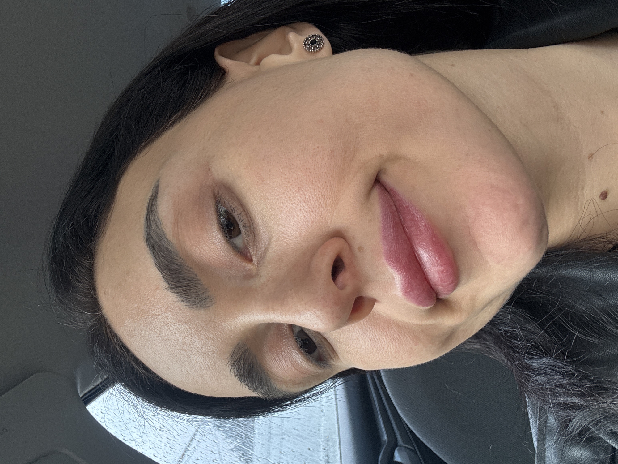 The image is a close-up selfie of a woman with dark hair and light skin. She has dark eyebrows and is wearing an earring in her left ear. Her lips are full and have a slightly glossy appearance.
The background is partially visible through the car window, and water droplets can be seen, indicating that it might be raining. The car's interior is also visible, with a black surface near the woman's shoulder.
There is no information in the image to indicate this photo was taken in Tbilisi, Georgia.