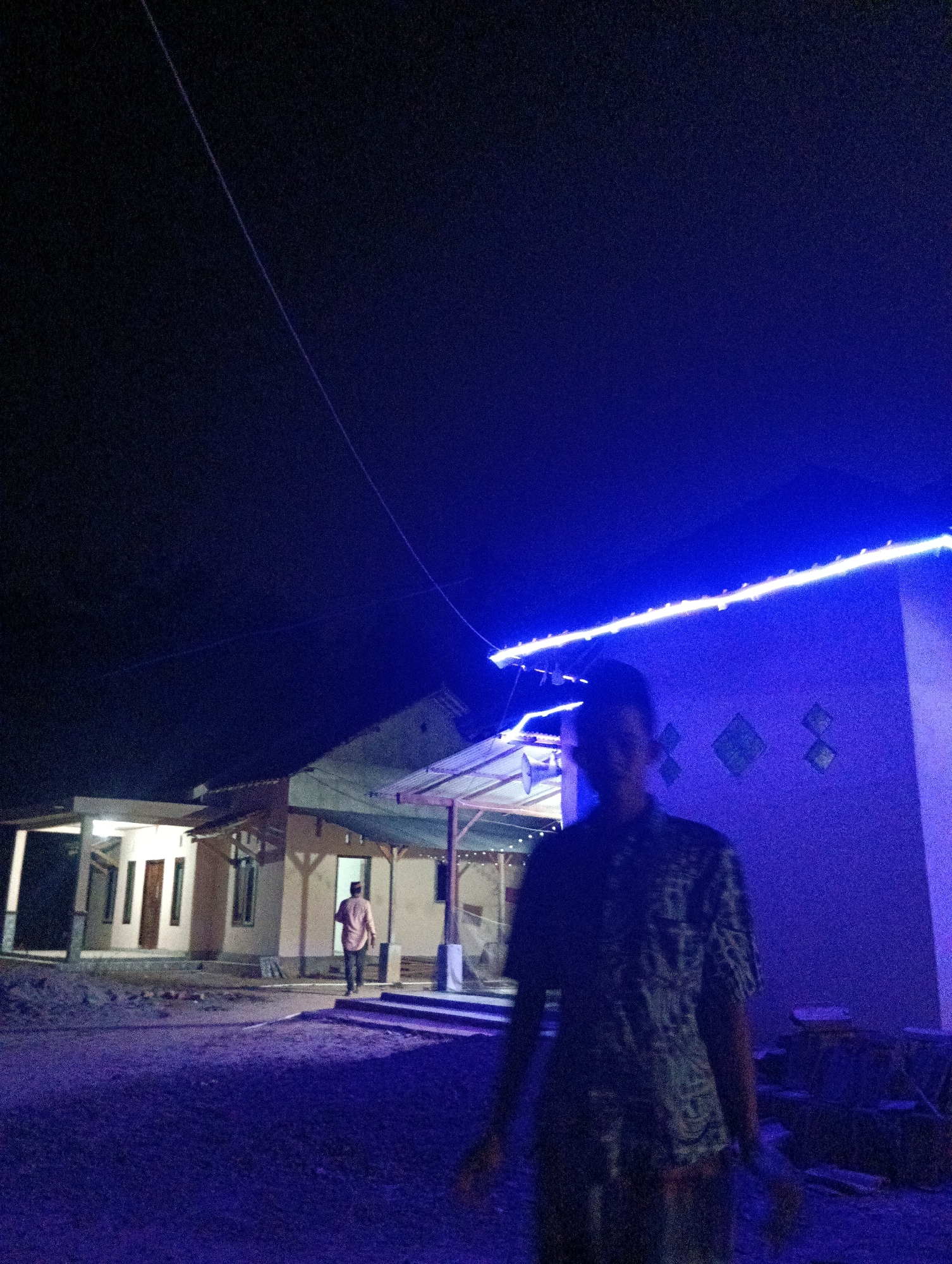 An outdoor, medium-shot, nighttime image captures a person in the foreground, partially obscuring a traditional Indonesian house. The person, a young male wearing a patterned shirt, stands to the right, facing slightly left. His features are indistinct due to low light and motion blur. Behind him, the house is illuminated by a vibrant blue LED light strip running along its roofline. The house appears to be a single-story structure with a covered porch. In the background, another individual, dressed in a light-colored shirt and dark pants, walks away from the camera towards the house. The ground is covered in gravel or dirt, and the overall atmosphere is dark, punctuated by the artificial blue light. The scene is likely set in Pematang Jaya, Indonesia, suggested by the architectural style and the mention of the location.