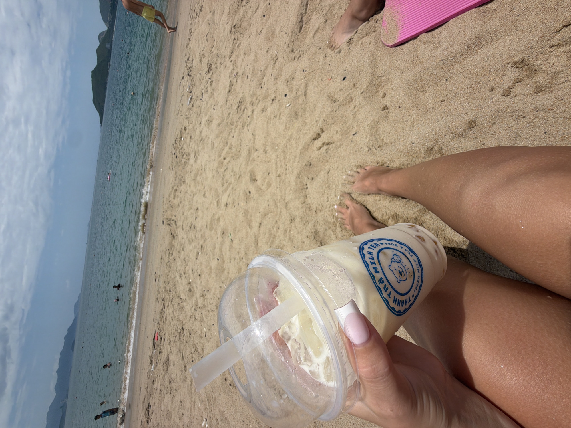 The image shows a close-up of a plastic cup of milk tea with boba, held by a person's hand with light pink painted fingernails. The cup has a blue and white label that reads "THANH TRA MILK TEA" and "FRESH MILK TEA & BOBA." The cup is placed on a sandy beach, with the legs of the person holding the drink also visible. In the background, the ocean stretches out to the horizon, with islands and hills visible in the distance. Several people are in the water, and one person in yellow shorts is walking on the beach. The sky is partly cloudy. The scene is consistent with a beach setting in Nha Trang, Vietnam, known for its beaches and coastal scenery.