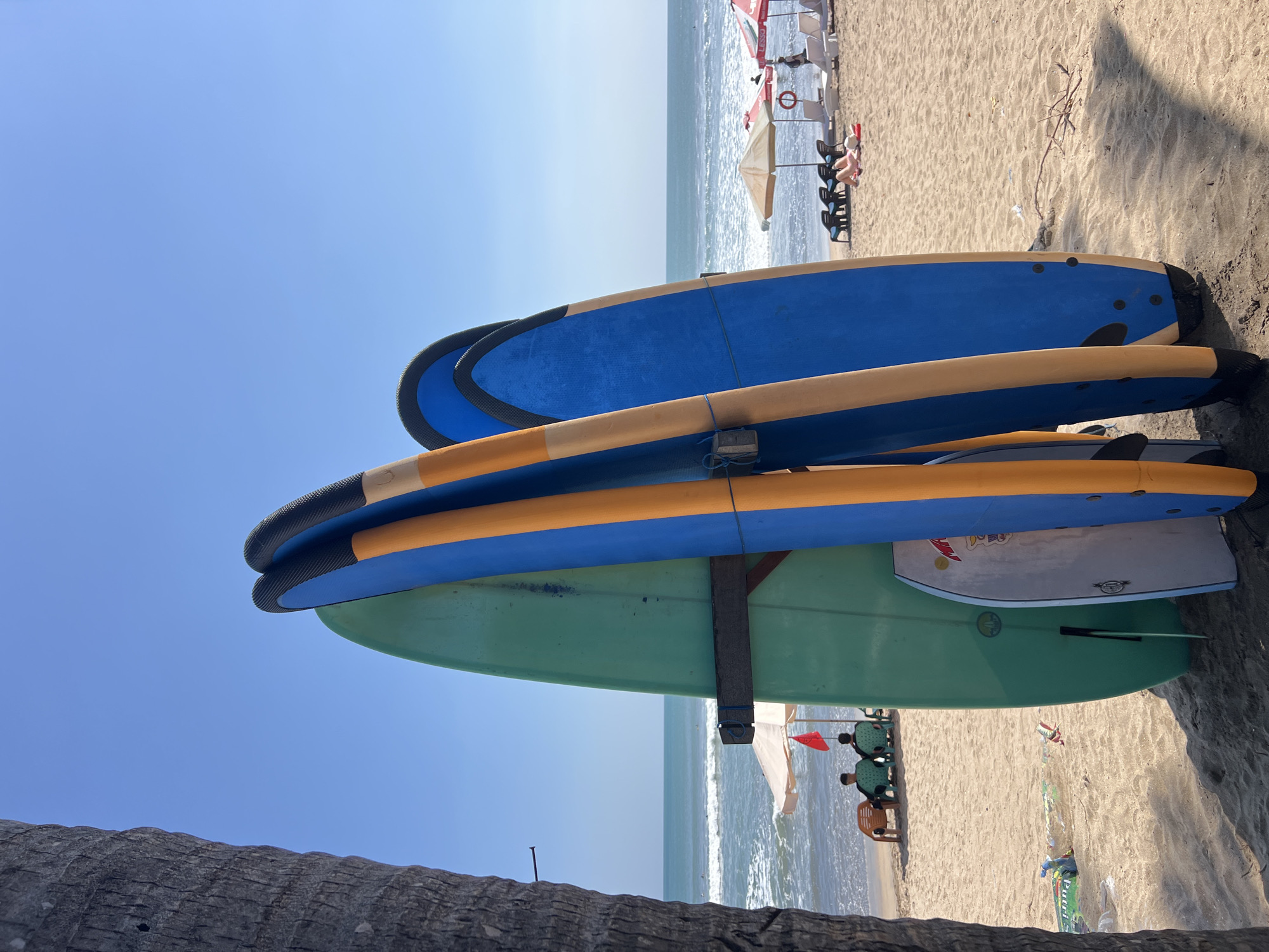 In Kuta, Indonesia, four surfboards are propped up on a sandy beach. The surfboards vary in color, with one being a bright green, another blue and orange, and a third primarily blue. To the left of the surfboards, a palm tree trunk is visible. Beyond the surfboards, the ocean stretches out under a clear blue sky. Waves are rolling in, and a few people can be seen in the water. Further down the beach, lounge chairs and umbrellas are set up, with some individuals relaxing. The scene depicts a typical day at a popular surf spot.