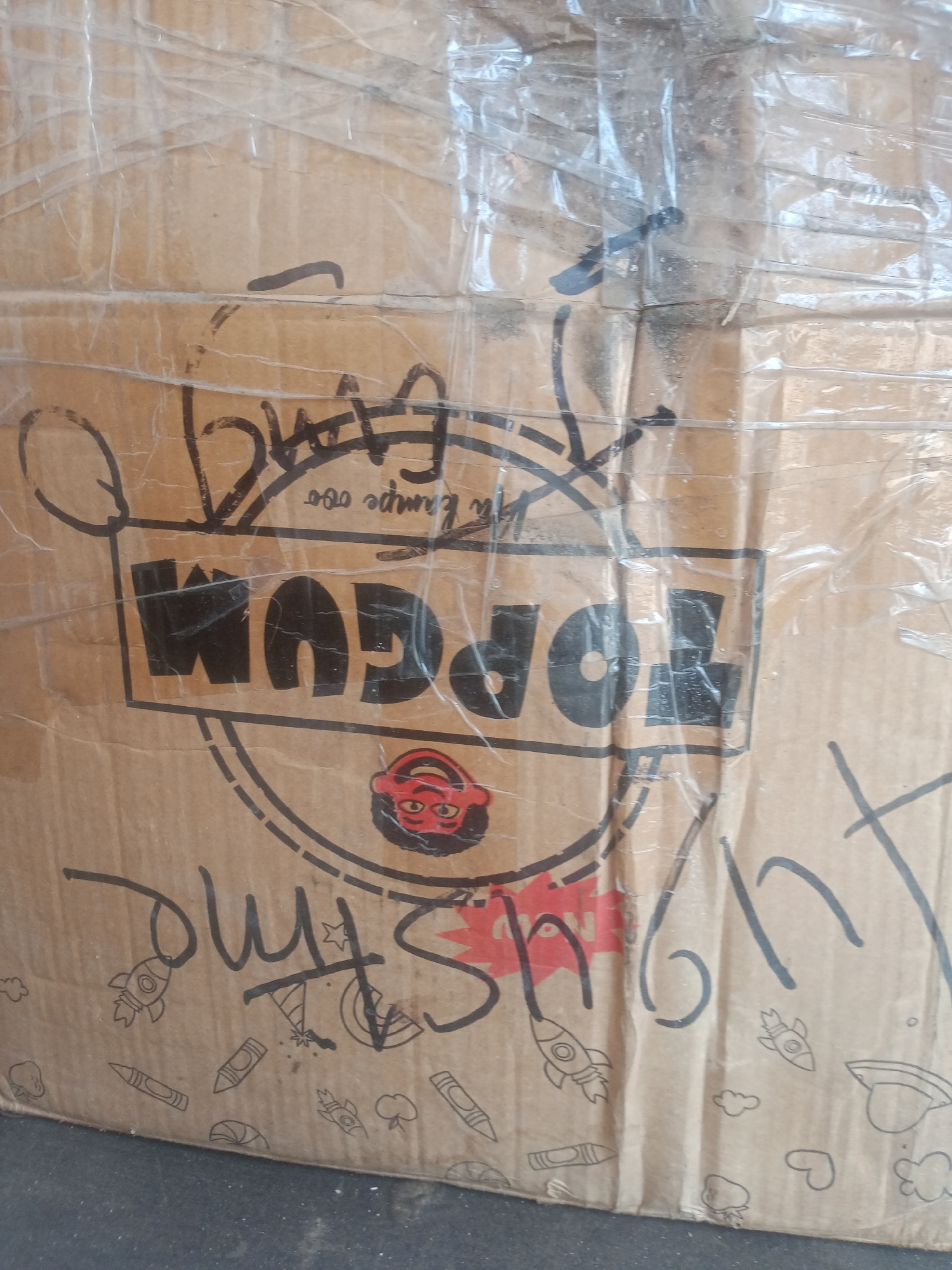 The image shows a close-up, eye-level view of a cardboard box. The box is taped with clear packing tape. The surface of the box has black marker writing, including what appears to be a logo or brand name "TOPGUM" in large, bold letters. There is also a drawing of a red-faced cartoon character with wide eyes and an open mouth. Below the "TOPGUM" text, there is writing that appears to be "it's time" or similar, along with various cartoon drawings of rockets, crayons, and party hats. In the upper portion of the image, there is more black marker scribbling. The background is indistinct, but appears to be a dark, possibly textured surface. The image does not contain any identifiable features that would allow for localization within Jalingo, Nigeria, or any other specific city.