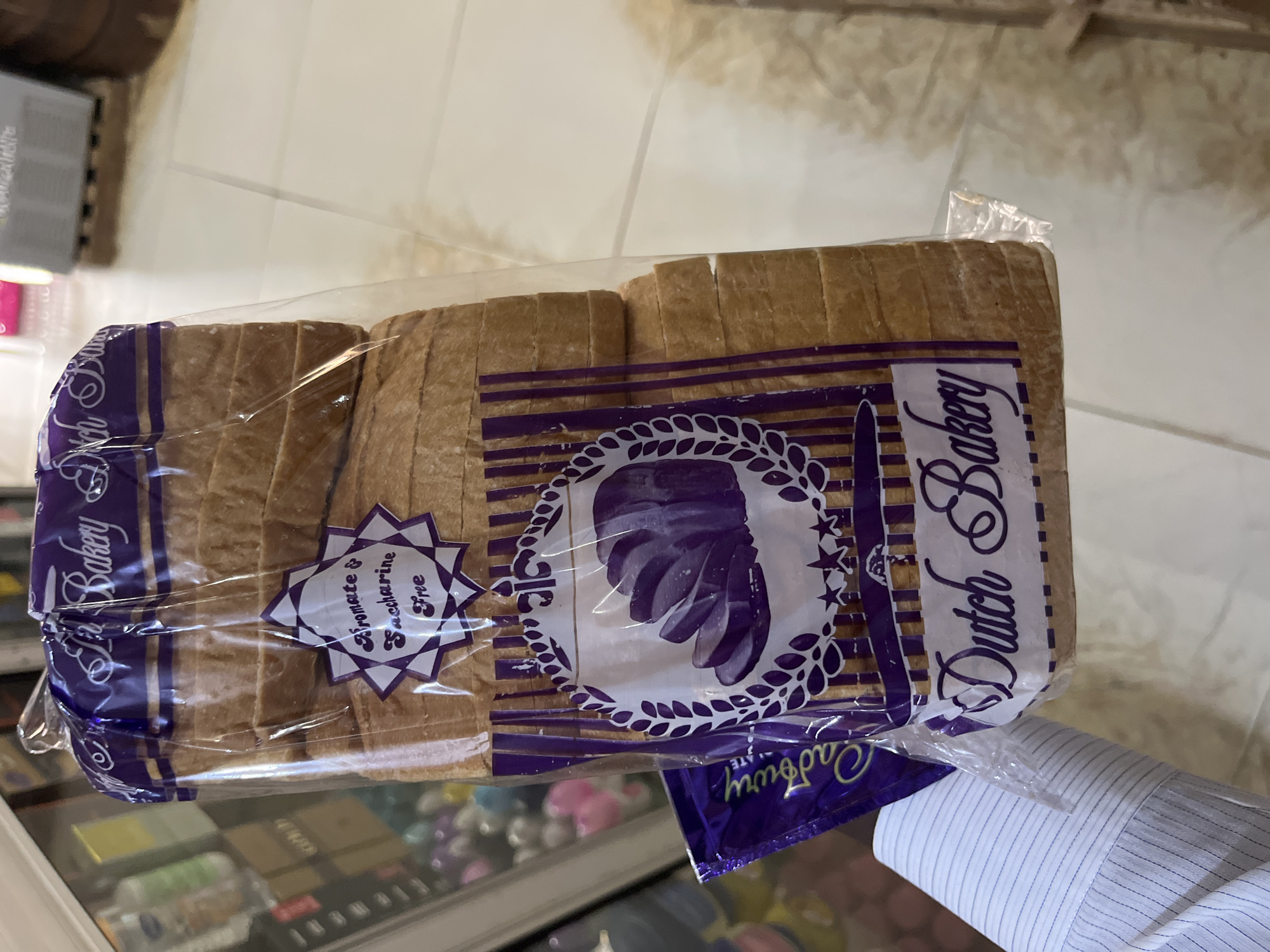 An indoor, close-up, eye-level shot shows a loaf of sliced white bread in a clear plastic bag. The loaf has a golden-brown crust and is neatly sliced. The plastic bag is printed with purple text and graphics. Prominently displayed is the text "Dutch Bakery" in a decorative script. There is also a star-shaped graphic with the words "Bromate & Saccharine Free" printed inside. In the lower left corner of the image, a small purple wrapper with "Cadbury" visible is tucked beside the bread. The background is blurred, but appears to be a retail environment with shelves and products. The lighting is bright.