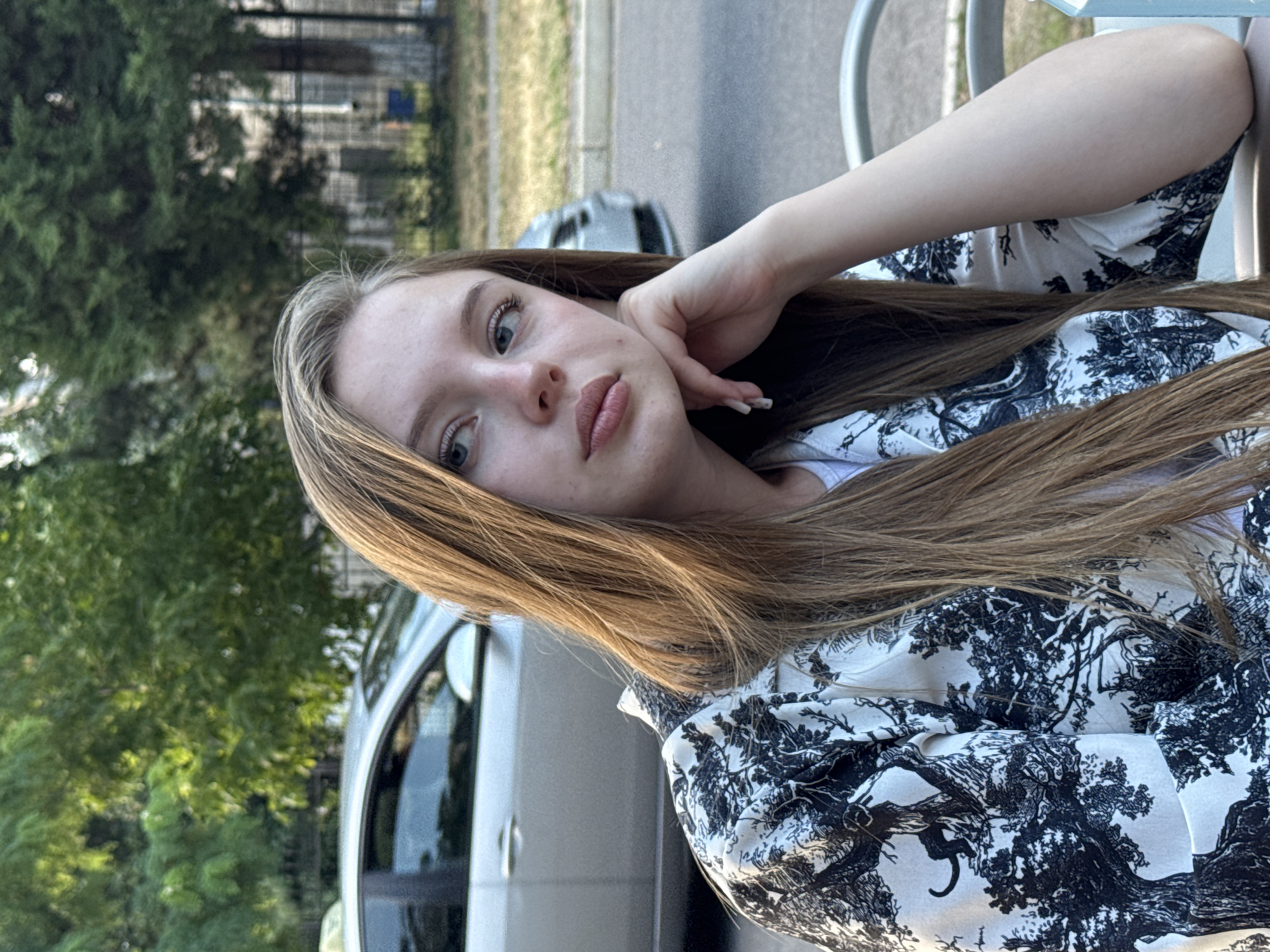 A young woman with long, light brown hair is shown from the chest up, resting her chin on her hand. She is wearing a white shirt with a black tree and animal print pattern. Her eyes are looking to her left. In the background, a silver car is parked on the street, and trees and a fence are visible. The scene appears to be outdoors in a city setting, possibly Tiraspol, Moldova, based on the presence of trees and a car on a street, common urban elements.