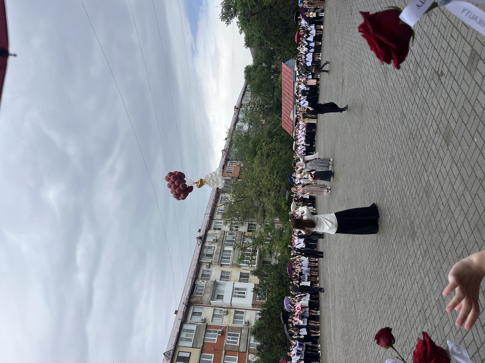 A low-angle, horizontal medium shot depicts a school graduation ceremony in Kaspiysk, Russia. Rows of students in black and white uniforms and graduation caps are lined up on a paved courtyard. Many hold roses. Some students hold red and white balloons. A medium-rise building forms the backdrop. Overhead wires stretch across the cloudy sky. A rose and part of a child's hand is visible in the foreground.