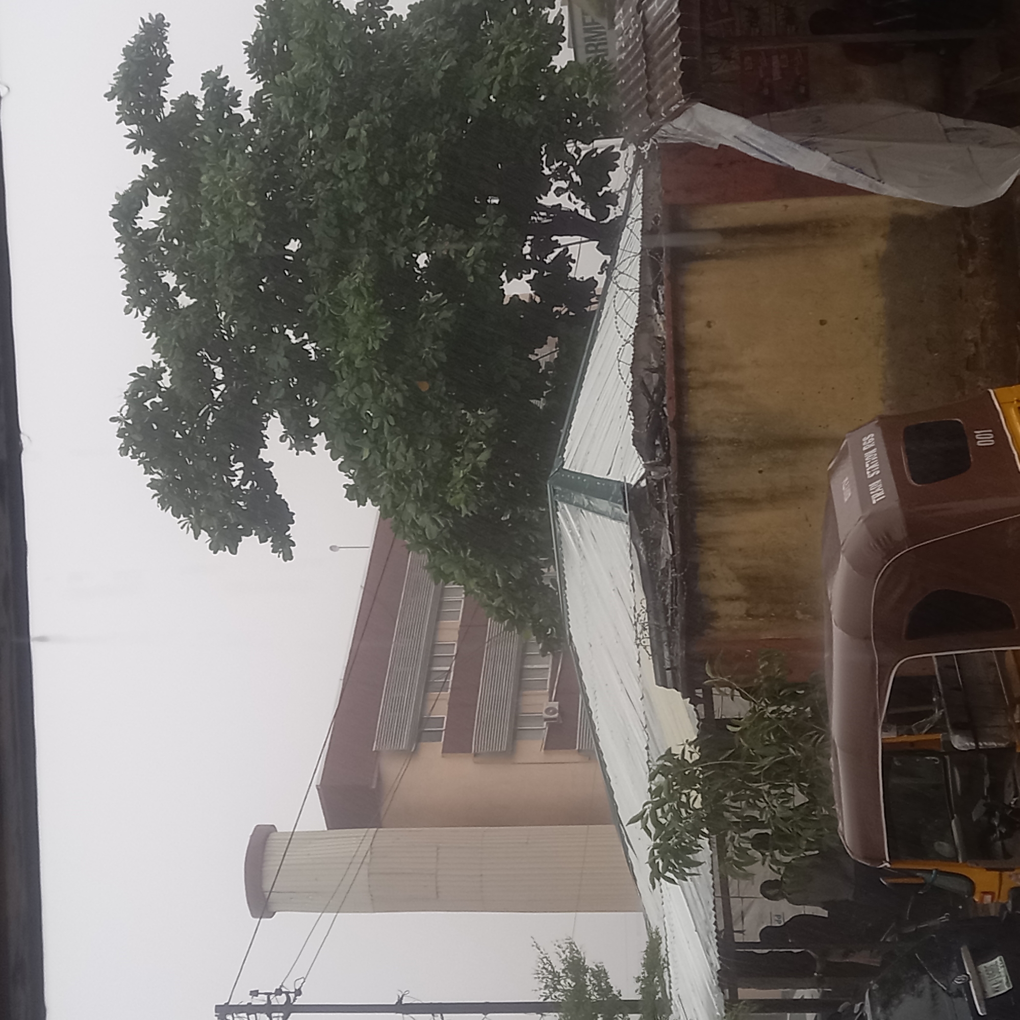 The image depicts a hazy outdoor scene, likely during rainfall, with limited visibility. In the background, a multi-story building with a brown facade and rows of windows with horizontal awnings is visible, alongside a tall, cylindrical structure with a light-colored ribbed exterior. A large, dense green tree dominates the mid-ground. In the foreground, a corrugated metal roof structure is visible, with barbed wire along its edge. A yellow and brown auto-rickshaw is parked in front of this structure, with part of a dark-colored vehicle to its left. Several figures, appearing to be people, are standing under or near the corrugated roof structure. The overall environment suggests an urban setting in Dadi Riba, Nigeria.