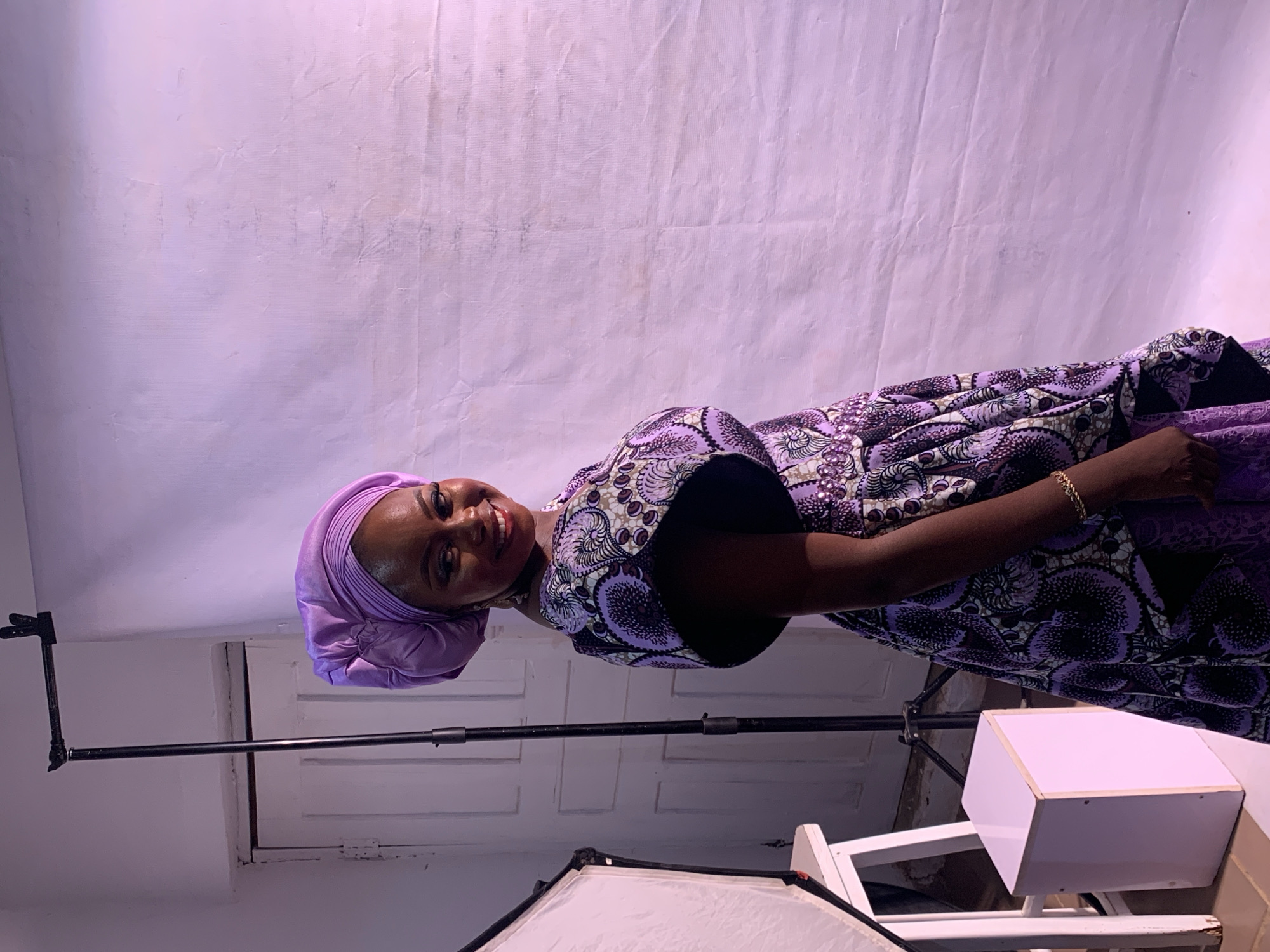 A young woman with brown skin and a purple head covering stands indoors in front of a textured white backdrop. She is wearing a patterned purple and black dress with large sleeves. She is smiling. A black metal support pole is visible behind her to the left. A white box and part of a photography softbox are visible to her right. The location is not specified as Jos, Nigeria in the image and cannot be determined.