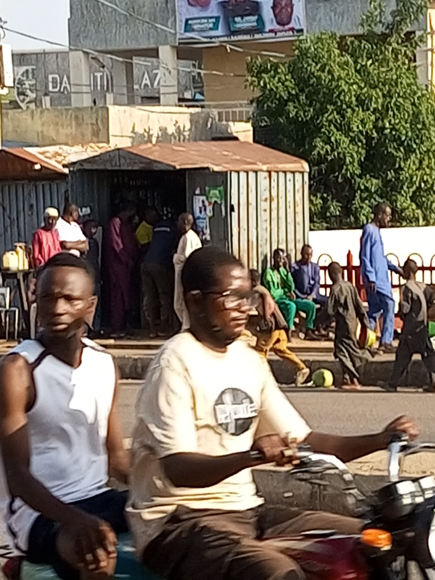 The image depicts a street scene in Tumfure, Nigeria. In the foreground, two men are seated on a motorcycle. The man on the left wears a white tank top and dark shorts, while the man on the right is in a light-colored t-shirt and brown pants. Both are looking forward. The motorcycle has a dark red body and a chrome handlebar with an orange indicator light.
In the middle ground, several people are standing or walking near a small, corrugated metal structure with a rusty roof. A man in a blue robe and trousers is walking from right to left, carrying a yellow object. To his left, another man in a dark garment is also walking, and a green watermelon lies on the ground nearby. Further left, a group of men are seated on a bench. One man is wearing a bright green outfit, and another is in a dark blue robe. Behind them, more people are gathered, some in traditional clothing like purple robes. To the far left, a vendor's stand with yellow containers is visible.
The background shows buildings, including a concrete structure with the partially visible text "DA ITI AZ" and what appears to be a large billboard or advertisement. A dense green tree is prominent on the right side of the frame. The overall environment suggests a bustling urban or semi-urban area with commercial activity. The lighting indicates it is daytime, with shadows cast on the ground.