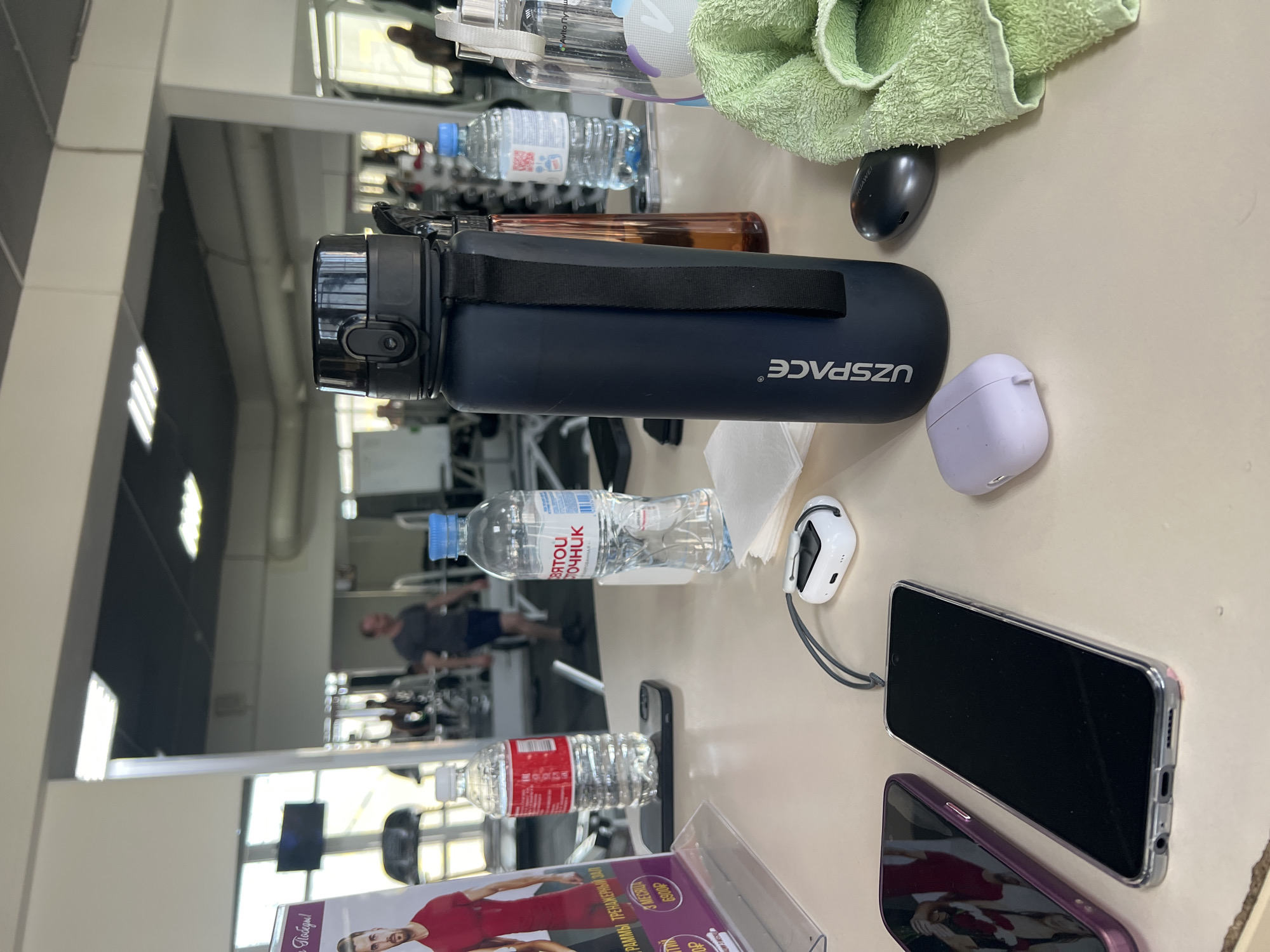 The image captures a snapshot from inside a gym or fitness center, with various personal items laid out on a light-colored table or counter. In the foreground, a dark blue "UZSPACE" brand water bottle with a black carrying strap is prominent. Beside it lies a light purple earbud case. A green towel is loosely placed near a small, dark, possibly Huawei-branded object.
Further along the table, a clear plastic water bottle with a blue cap and "СВЯТОЙ ИСТОЧНИК" (Svyatoy Istochnik) branding is visible, along with another clear bottle with a white cap and red label. A white earbud case with a gray lanyard is positioned next to a black-screened smartphone in a clear case. Another smartphone with a purple case reflects a man in a red shirt from a nearby promotional stand. This stand also displays text like "Победы!", "ГРАММЫ ТРЕНАЖЕРНЫЙ ЗАЛ" (likely "Gym Programs"), and "6900Р," indicating a cost in rubles. A brown, cylindrical bottle, possibly for fragrance or supplements, is also present.
The background, primarily composed of large mirrors, reflects the gym's interior, showing equipment and a man in a dark t-shirt and shorts engaged in exercise. The reflections suggest an active gym environment during what appears to be a break from a workout, with personal belongings temporarily stored on the side. The overall lighting suggests an indoor setting, likely during daylight hours given some light sources.