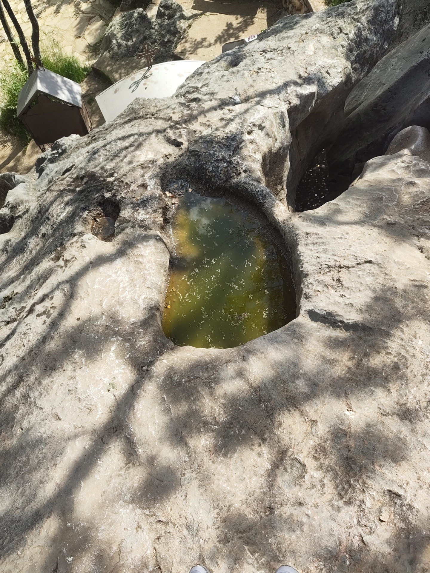 A high-angle shot shows a natural rock formation filled with greenish water, next to another cavity containing coins, likely a wishing well. The location is Starozagorski bani, Bulgaria, as deduced from associated information. A corner of what appears to be a white structure and a donation box are visible at the top left. Shadowy tree limbs cover the rock surface.