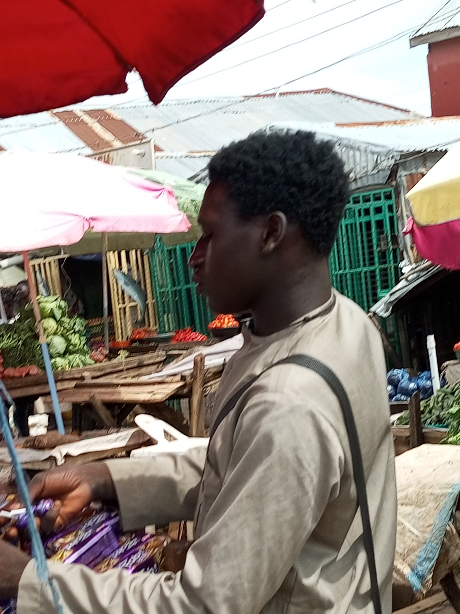 The image captures a side profile of a young Black man in a marketplace in Tempure, Nigeria. He wears a light brown, collared shirt with a dark strap across his chest, suggesting he might be carrying a bag. His short, dark, curly hair frames his face. His right hand is engaged in handling items that appear to be wrapped snacks or candies, possibly in purple packaging. He is standing behind a table or stall. In the background, the market is visible with stalls covered by various colored tarpaulins, including red, pink, and yellow. Shelves and structures made of wood and corrugated metal are present, and goods like leafy vegetables and what seem to be red fruits or tomatoes are displayed. A suspended fish can be seen in one of the stalls, indicating a food market. The overall scene depicts a typical bustling market environment.