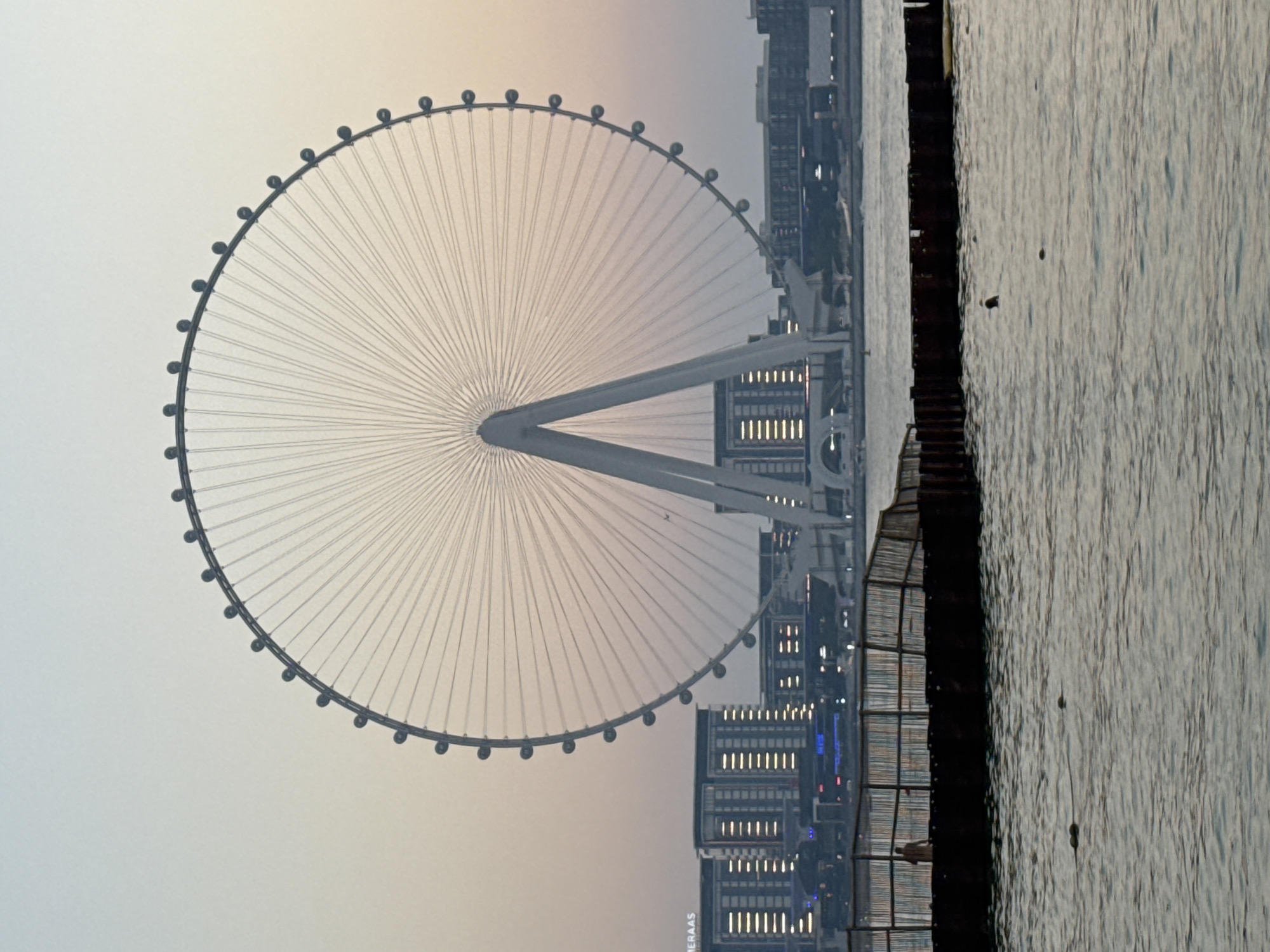 The image shows the Ain Dubai observation wheel in the city of Dubai, United Arab Emirates, at dusk. The massive Ferris wheel dominates the frame, with its spokes radiating outward from a central structure. The sky is a gradient of pale blue and soft pink, indicating the transition from day to night. In the background, modern buildings with illuminated windows form a cityscape. In the foreground, calm water stretches across the bottom of the image. A temporary-looking structure made of wood and metal, possibly a pier or a barrier, is visible in the middle ground, with a single person standing on it. Several buoys float on the water's surface. The overall scene depicts a serene evening view of a prominent Dubai landmark and its surrounding urban and natural environment.