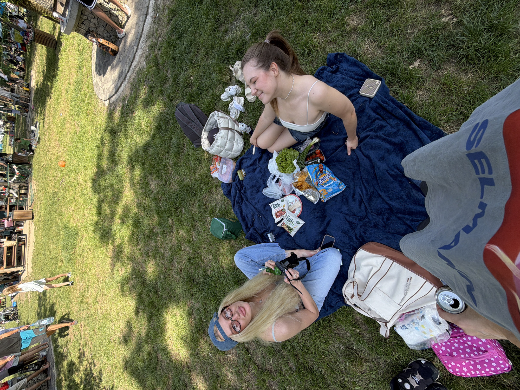 A high-angle medium shot captures two young women enjoying a picnic on a sunny day in Cukarica Urban Municipality, Serbia. They are seated on a dark blue blanket spread on a grassy area. One woman with blonde hair and sunglasses is sitting cross-legged, holding a small bottle and camera. The other woman, with brown hair in a ponytail, is kneeling and looking off to the side. A variety of items are spread around them, including snacks, a bag of chips, lettuce, a small speaker, shoes, and bags. To the right of the frame, the lower leg and shorts of another person can be seen, holding a can of soda. In the background, there is a view of a public area with trees and people.