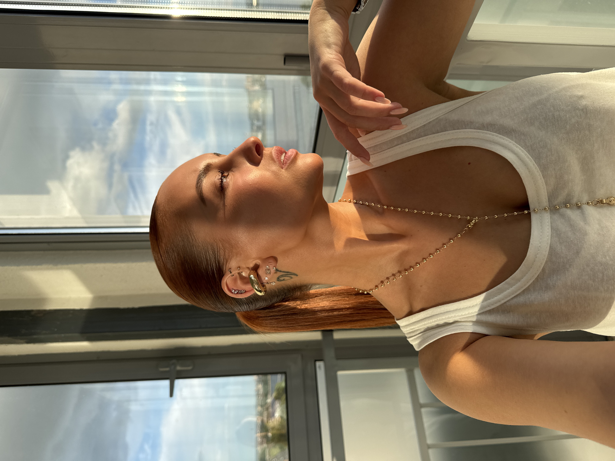 A person, possibly female, with reddish-brown hair pulled back in a ponytail and tanned skin, is seen in a sunlit indoor setting, likely a high-rise building or apartment, as suggested by the large window showing a cityscape. She is wearing a white tank top and a gold necklace. There is jewelry in the person's ear and a small tattoo is seen on her neck. The woman is looking upwards and has a hand near her neck. Based on the view through the window and the urban setting, it's possible the location is Izhevsk, Russia, though specific landmarks cannot be identified from the image.