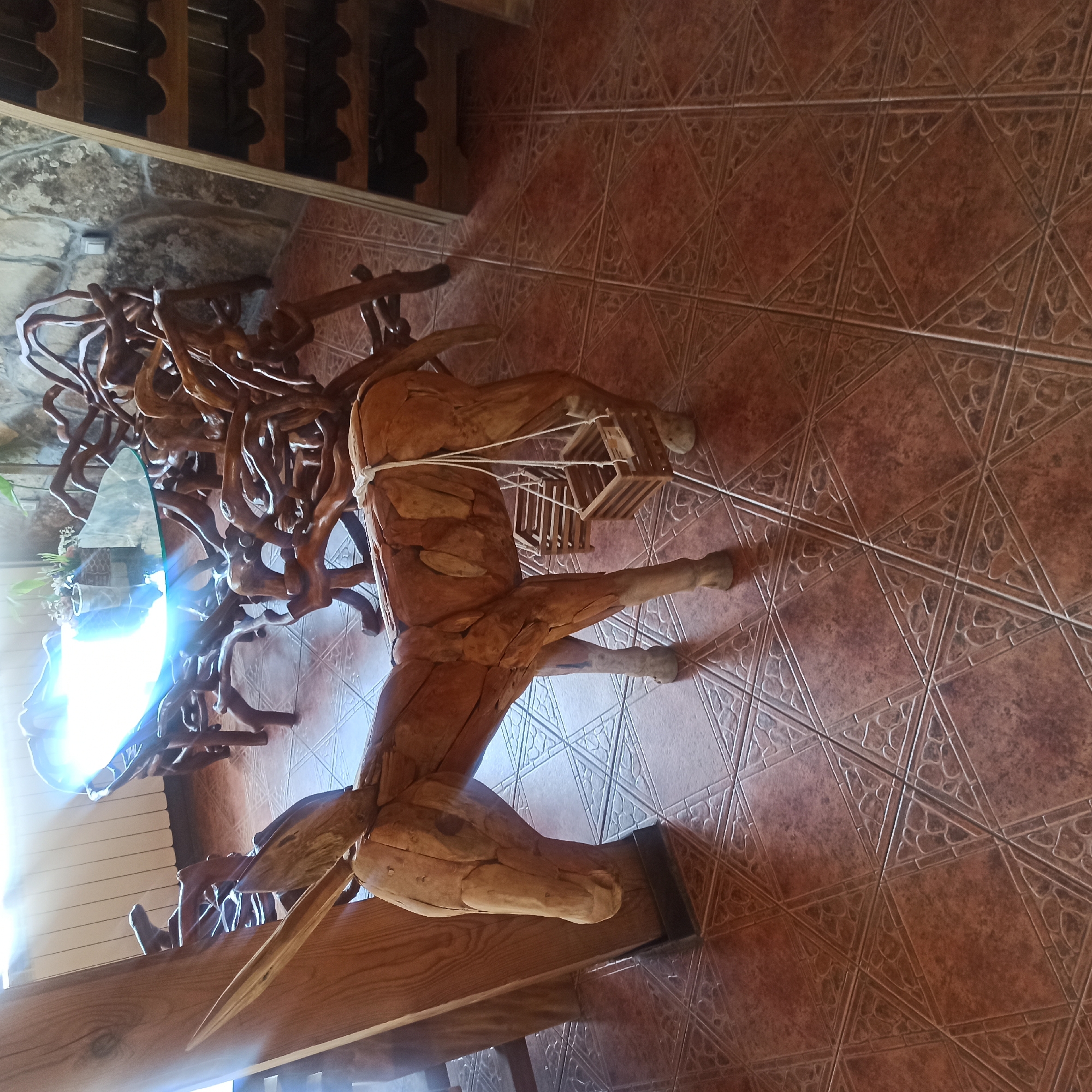 The image depicts a wooden sculpture of a donkey on a tiled floor. The donkey is positioned in the foreground, facing left, with two small wooden baskets hanging from its sides. Behind the donkey, there is a wooden table with a glass top, supported by an intricate base made of branches. In the background, a stone wall is visible, and to the right, a wooden wine rack filled with bottles. Sunlight streams in from the left, casting a bright glare on the table and the wall. The scene is indoors, likely in a restaurant or a rustic-themed establishment, given the presence of the wine rack and the natural wood elements. The overall aesthetic is rustic and artistic. This setting is consistent with the rural area of Zlatna Panega, Bulgaria, known for its natural beauty and potentially for establishments that embrace a natural, artistic decor.