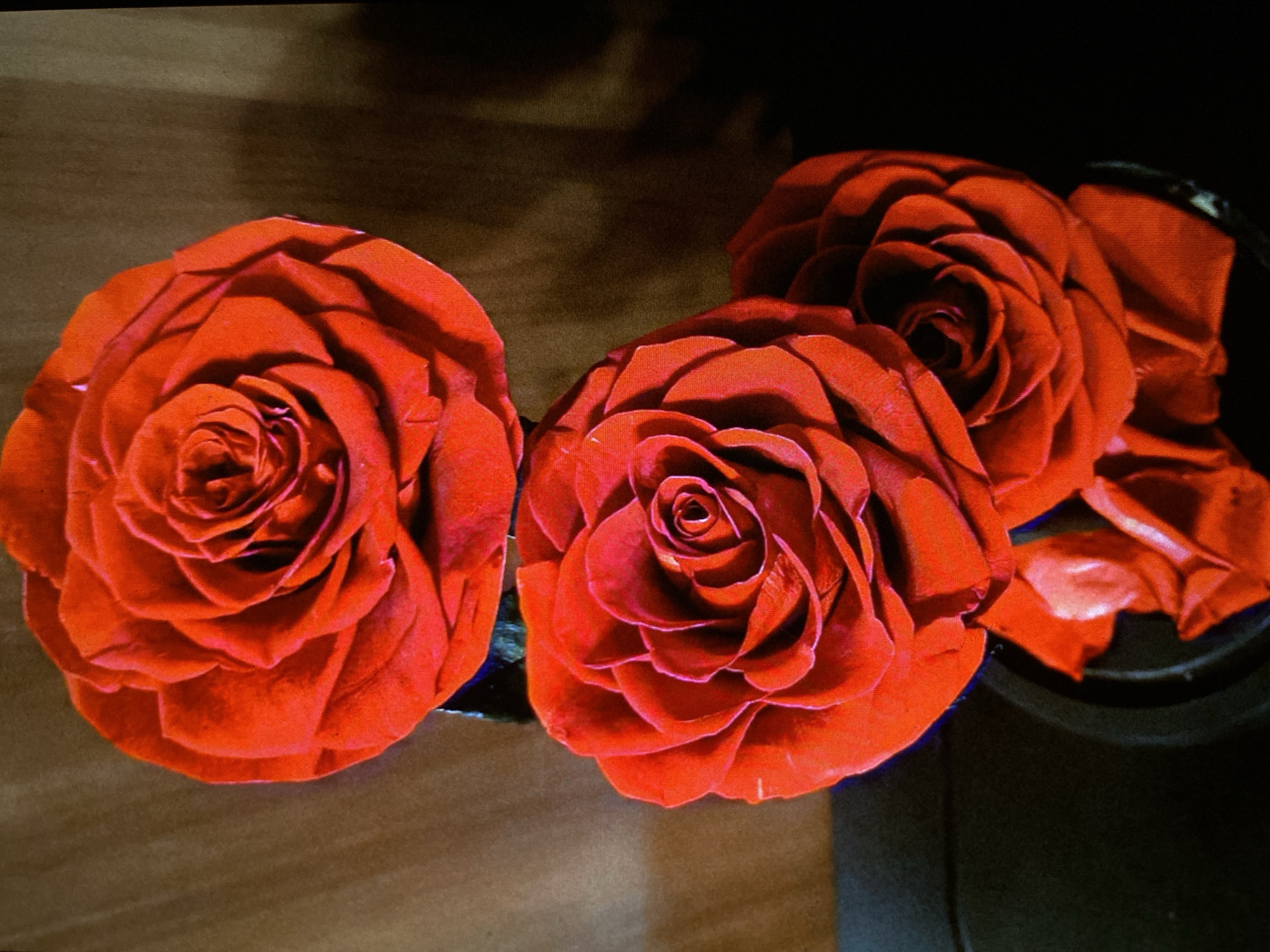The image displays a close-up, high-angle shot of three vibrant red roses arranged on a surface. The roses appear to be artificial, likely made of fabric or plastic, characterized by their uniform texture and striking, almost luminous red color. Two roses dominate the left and center foreground, each with intricately layered petals forming a central spiral. A third rose is partially visible behind and to the right, seemingly resting in a dark, bowl-like container or base.
The roses are illuminated, creating highlights on their petals and casting subtle shadows. The surface beneath the roses varies: a lighter brown, possibly wooden, texture is visible on the left and bottom, while the right side and background consist of darker, blurred areas, suggesting shadows or an indistinct background. There is no human activity depicted, nor any visible text. The time of day and weather are not discernible from the indoor setting. The scene is located in Bankya, Bulgaria, as per the provided context.